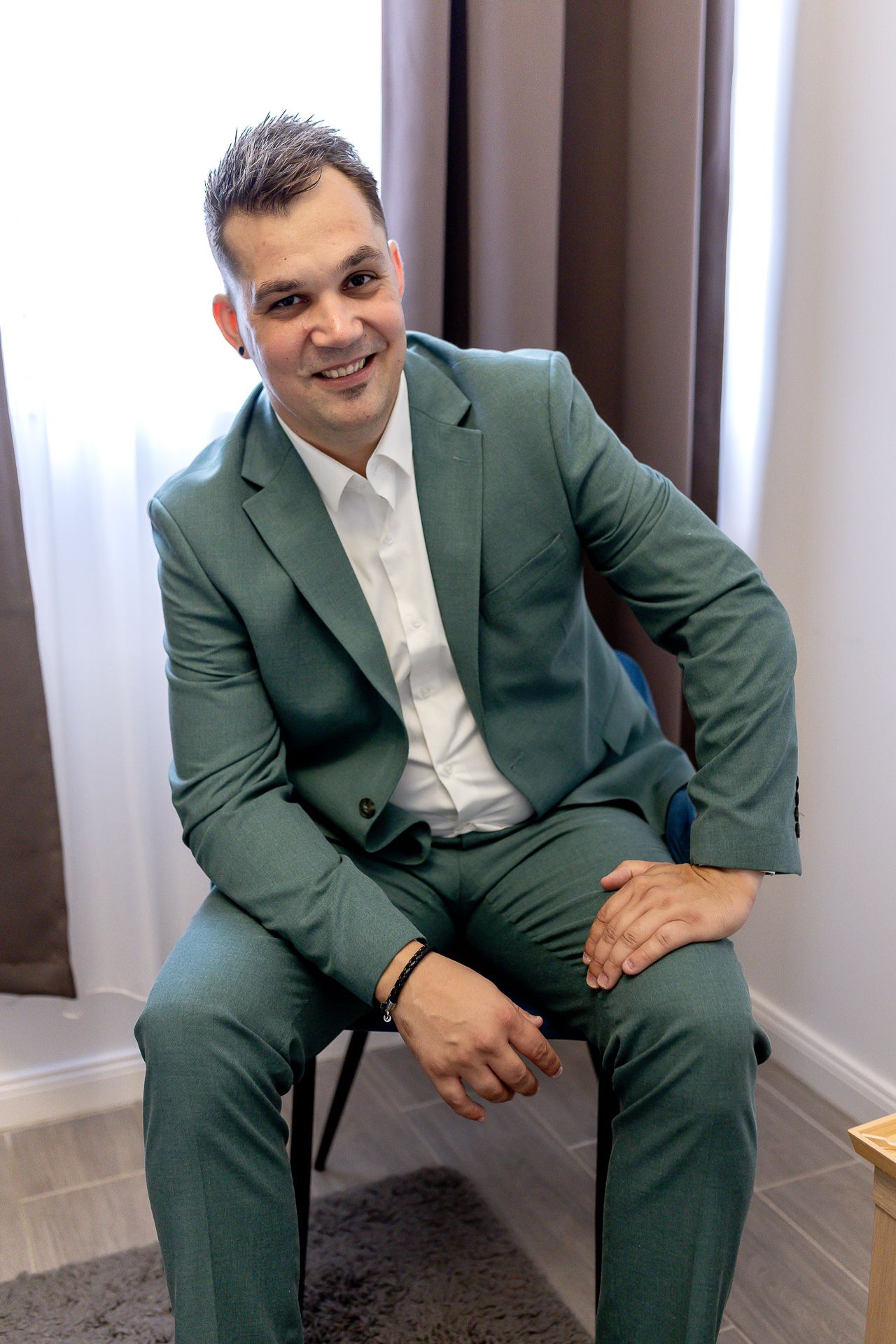 A person in a green suit and white shirt sits casually on a chair against a light-colored wall.