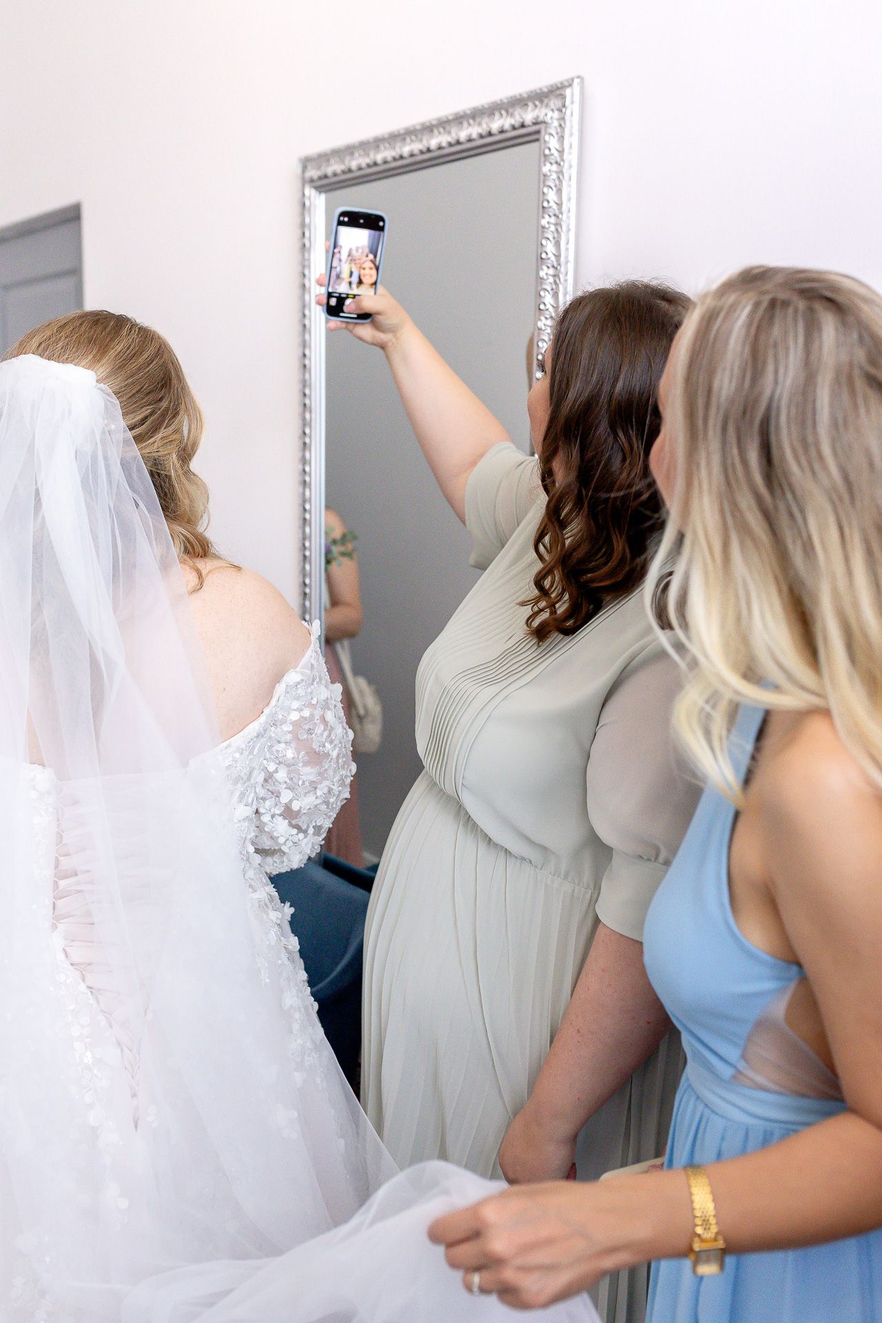 A group in formal attire gathers around a mirror, with someone wearing a flowing white veil in the center.