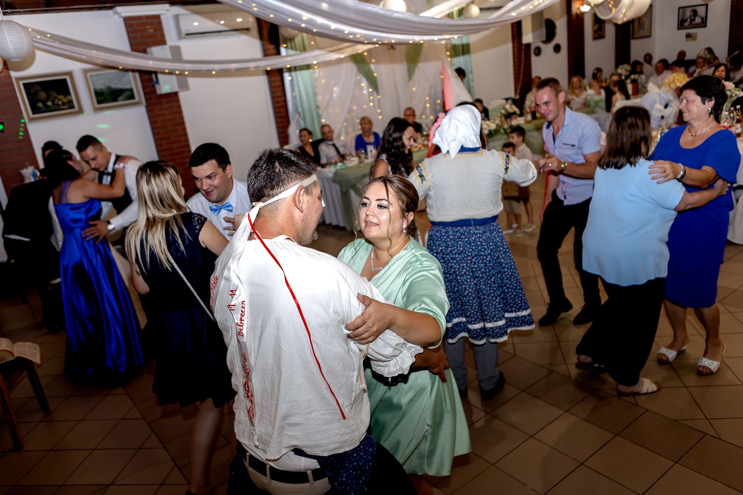 Couples dance together at a wedding reception in a decorated indoor venue with string lights overhead.