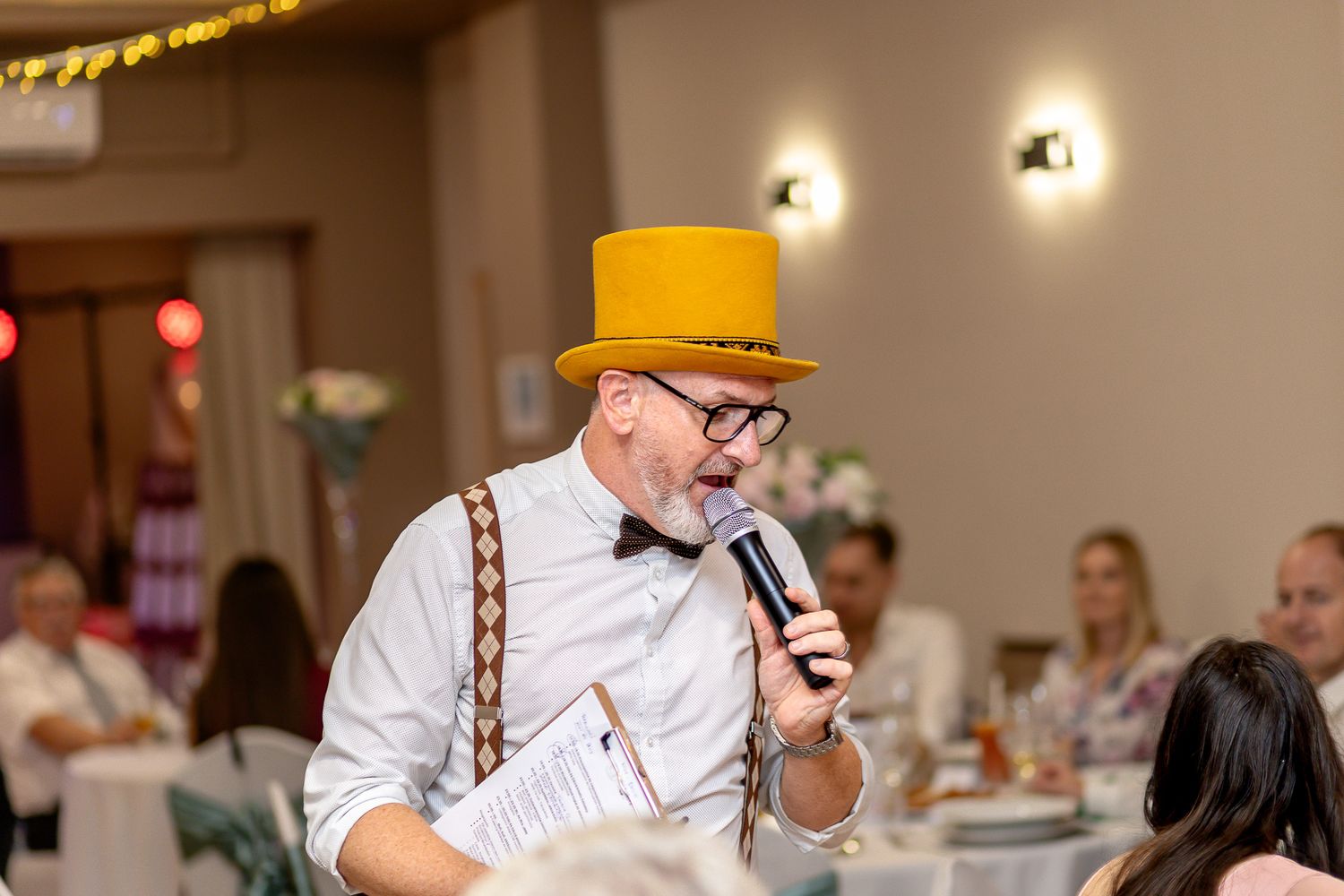 A speaker wearing suspenders and a yellow top hat addresses guests at an indoor event with wall sconces visible.