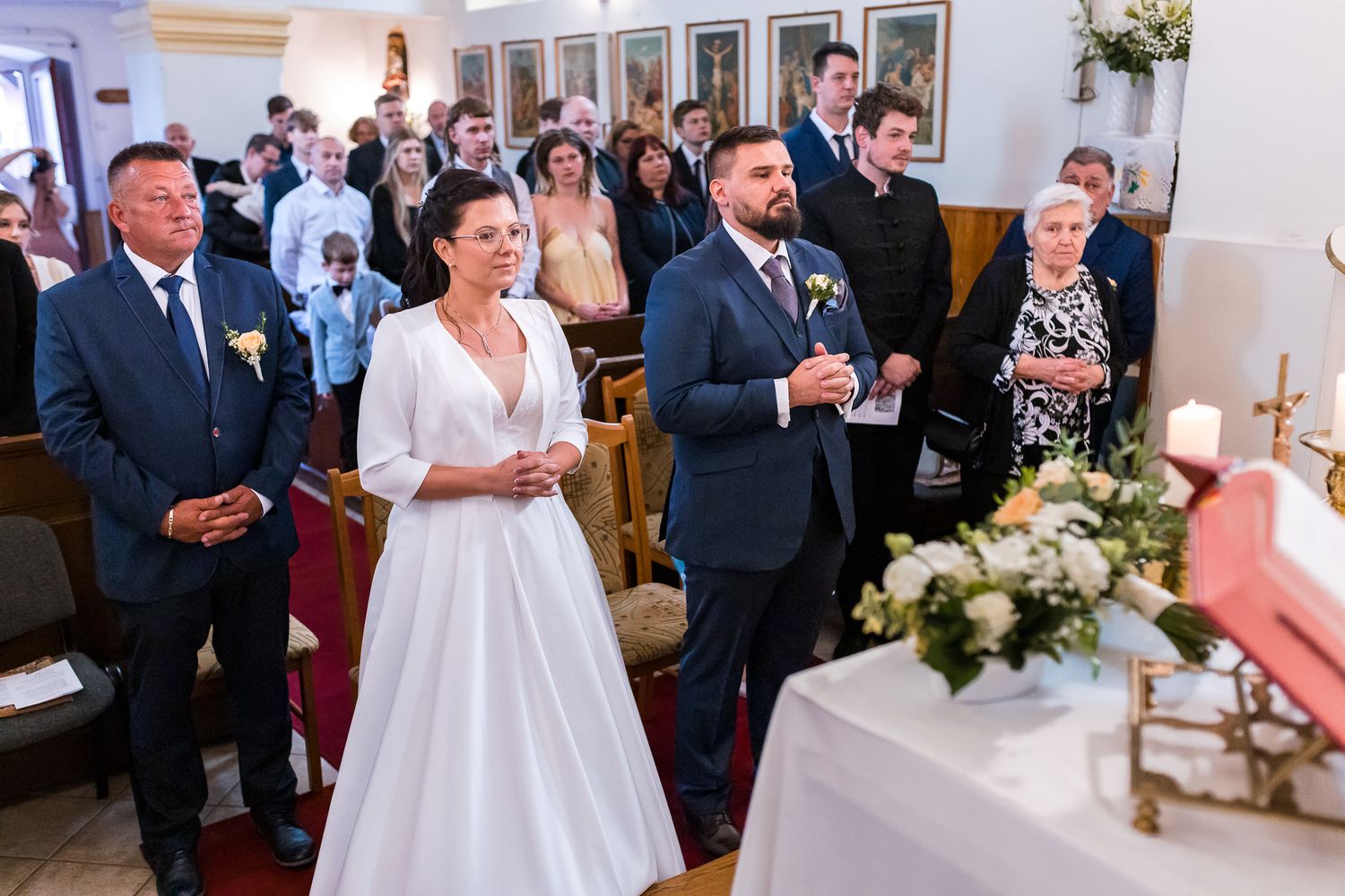 Wedding ceremony guests stand in a church during a traditional service with white floral decorations and candles.