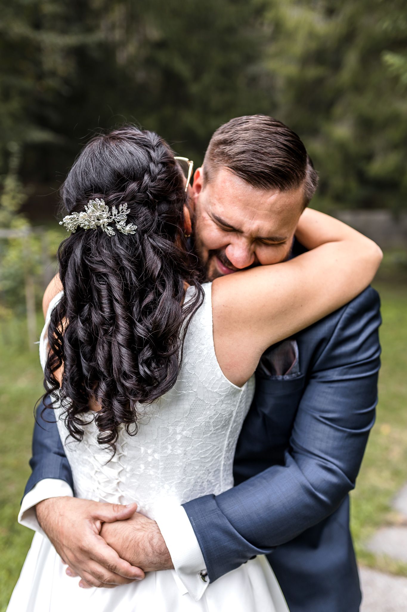 A romantic wedding couple embraces tenderly in an outdoor setting with natural greenery in the background.