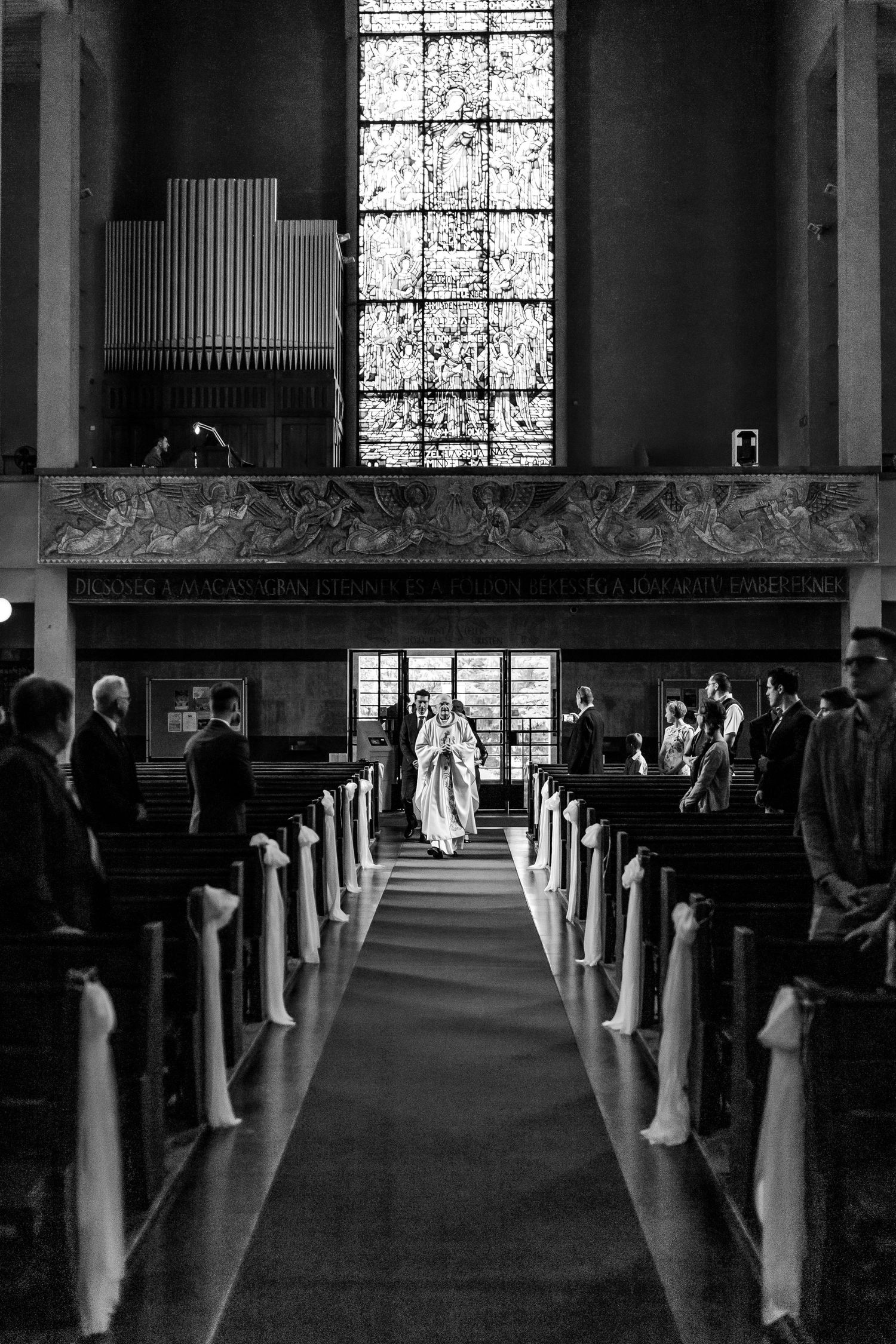 Black and white view of a church aisle lined with white decorative elements and stained glass windows.