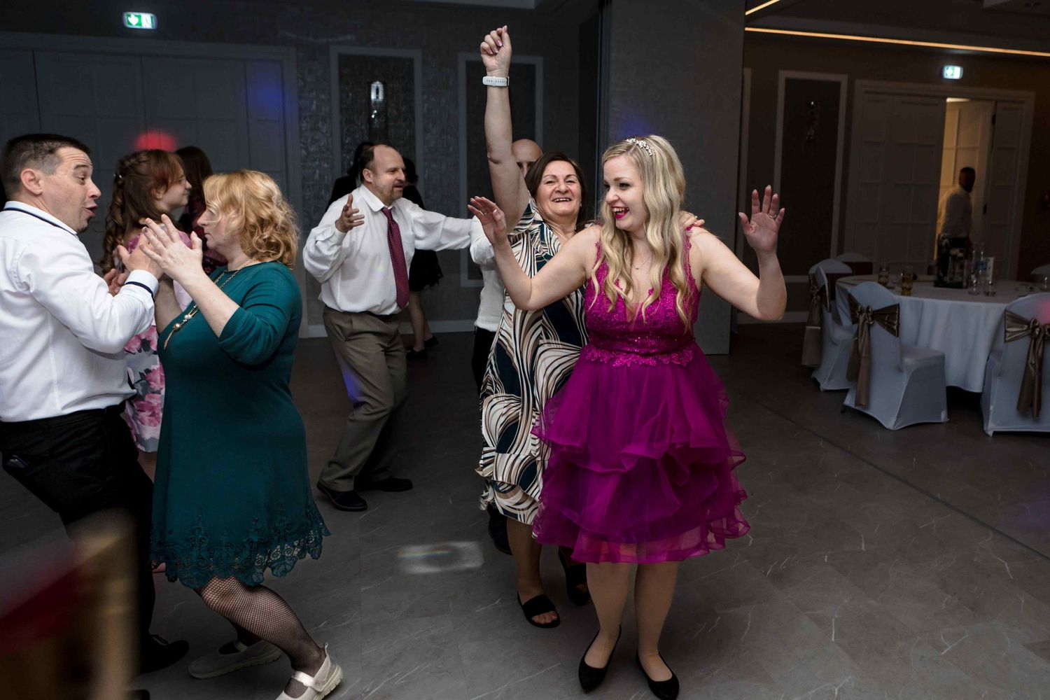 People dance and socialize in a dimly lit indoor venue with white chairs visible in the background.