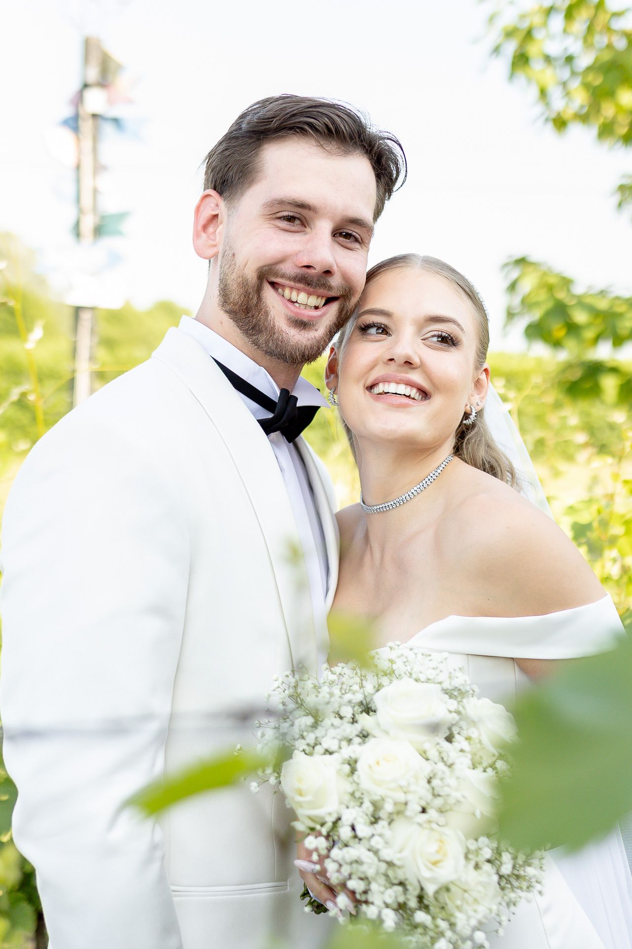 Romantic wedding couple embraces outdoors with bride holding baby's breath bouquet on sunny day.