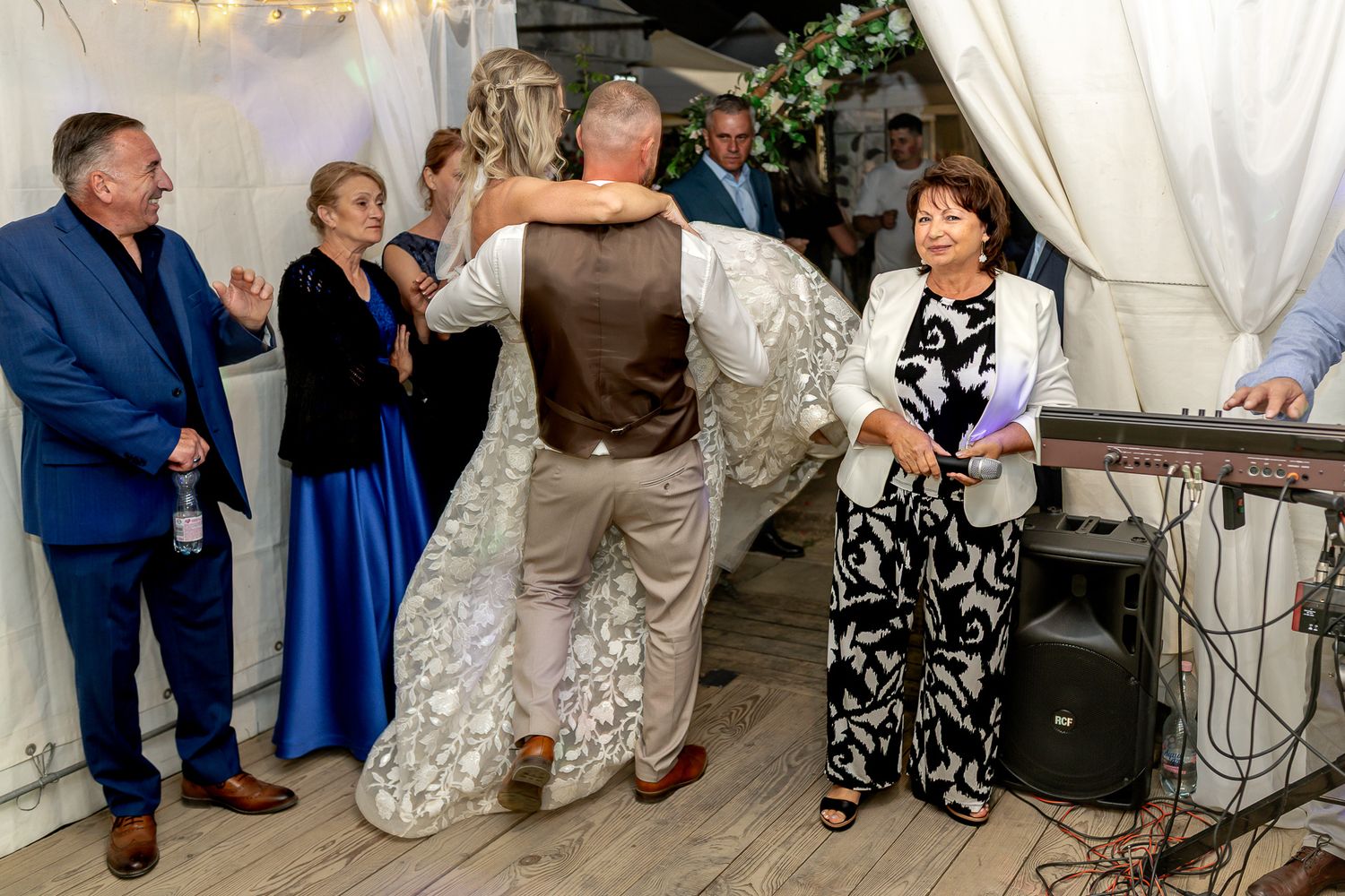 Wedding guests dancing and celebrating on a dance floor under elegant white drapes and chandeliers at an indoor reception.