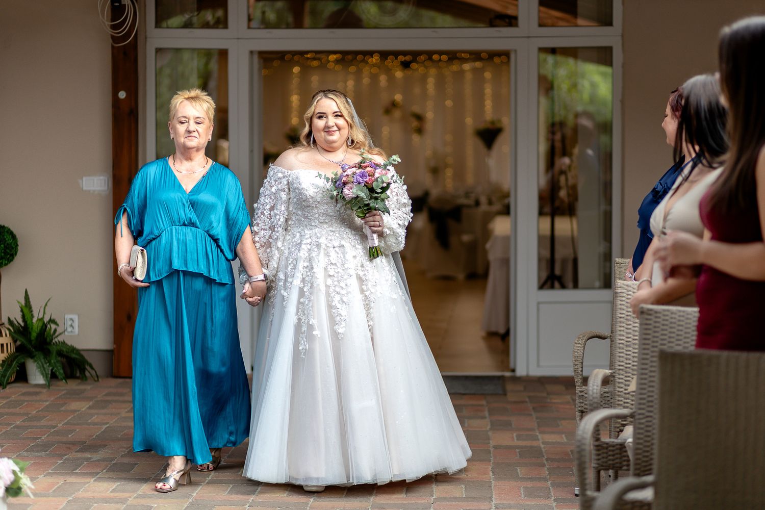 A person in a teal dress stands beside someone wearing a white wedding gown holding a floral bouquet outside a venue.