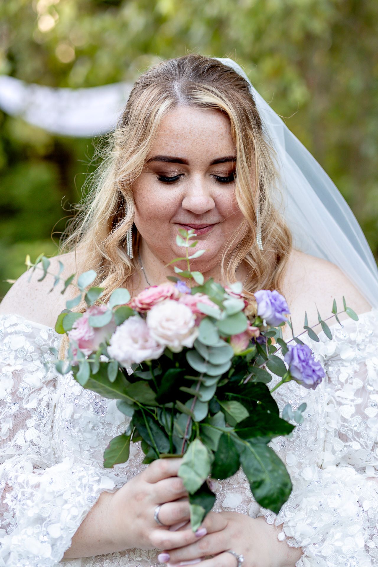 A bride in a white lace dress holds a romantic bouquet of pink roses and eucalyptus against natural outdoor lighting.
