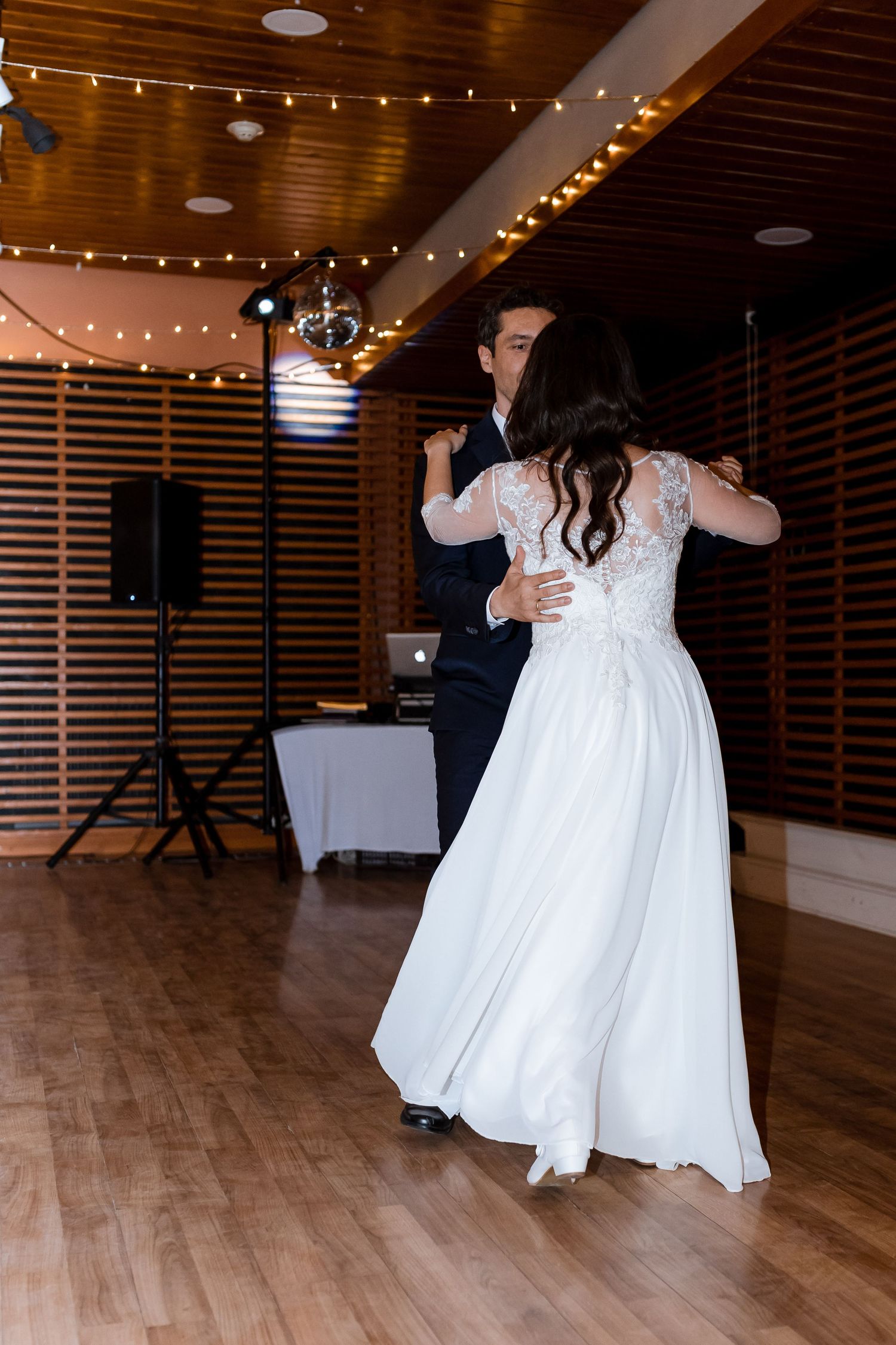 Two people in white wedding dresses dance together on a wooden floor under string lights at a reception venue.