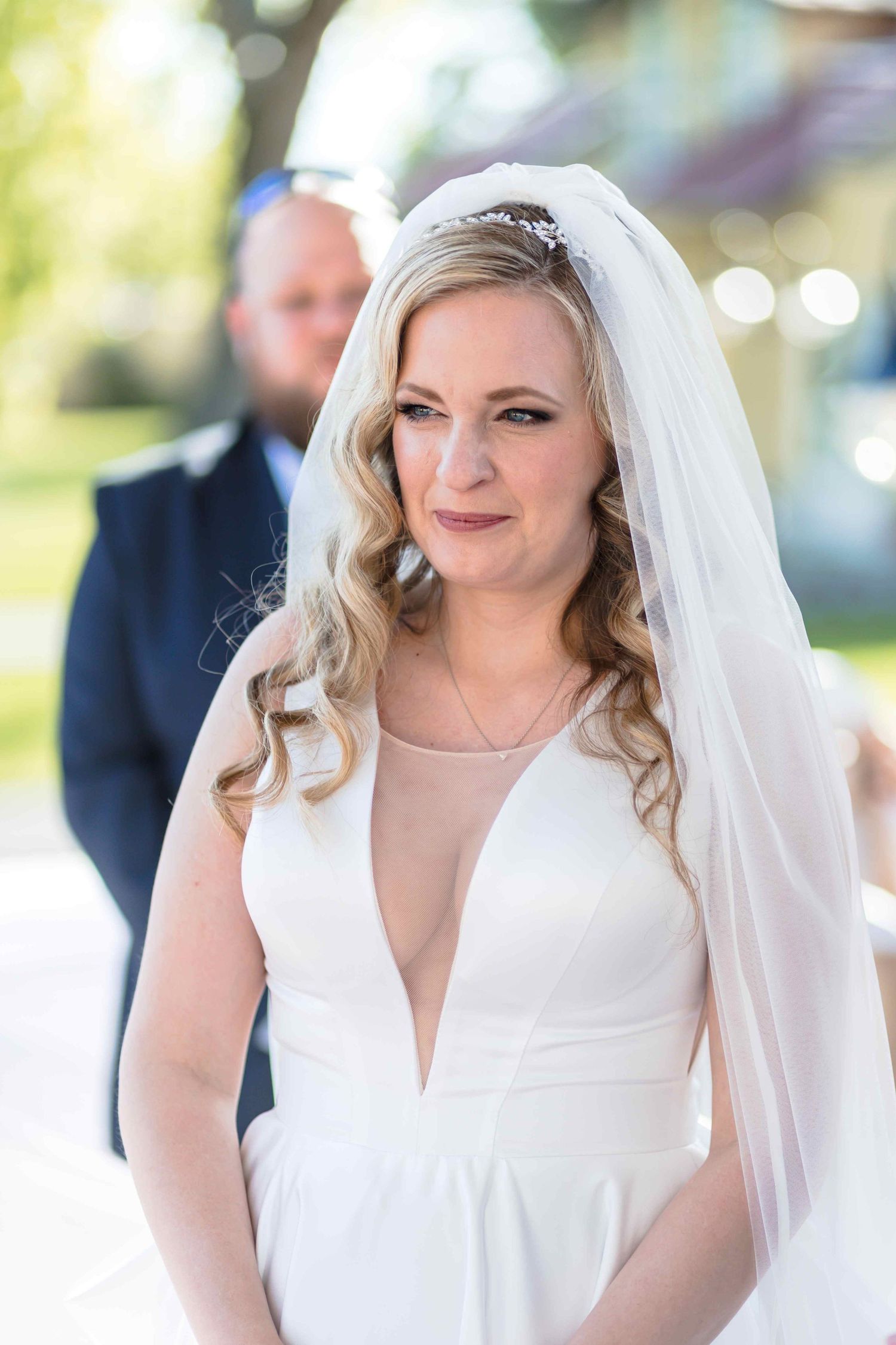 A bride in a white wedding dress and veil smiles joyfully during an outdoor wedding ceremony.