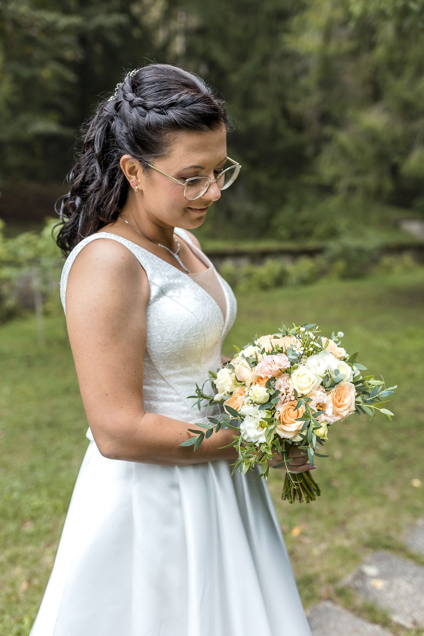 A bride in a white dress smiles while holding a peach and cream floral bouquet in an outdoor garden setting.