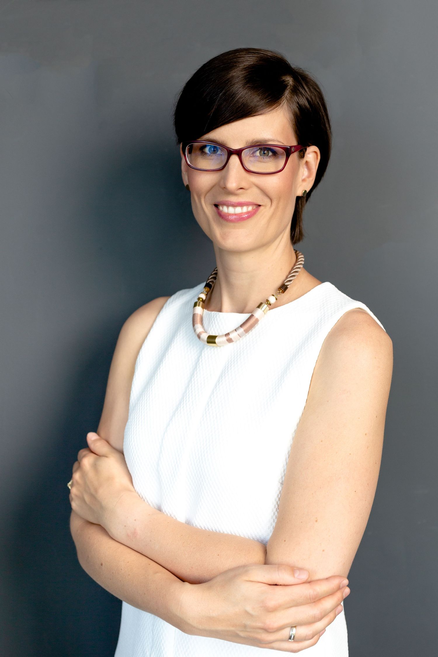 Professional headshot against grey backdrop showing confident pose with gold statement necklace.