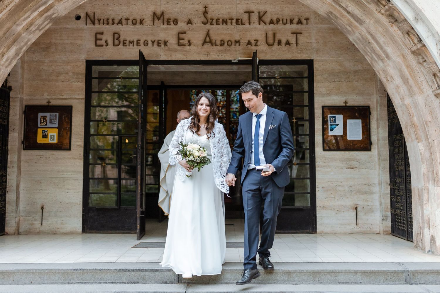 Bride and groom walk together under stone archways in an elegant outdoor setting.