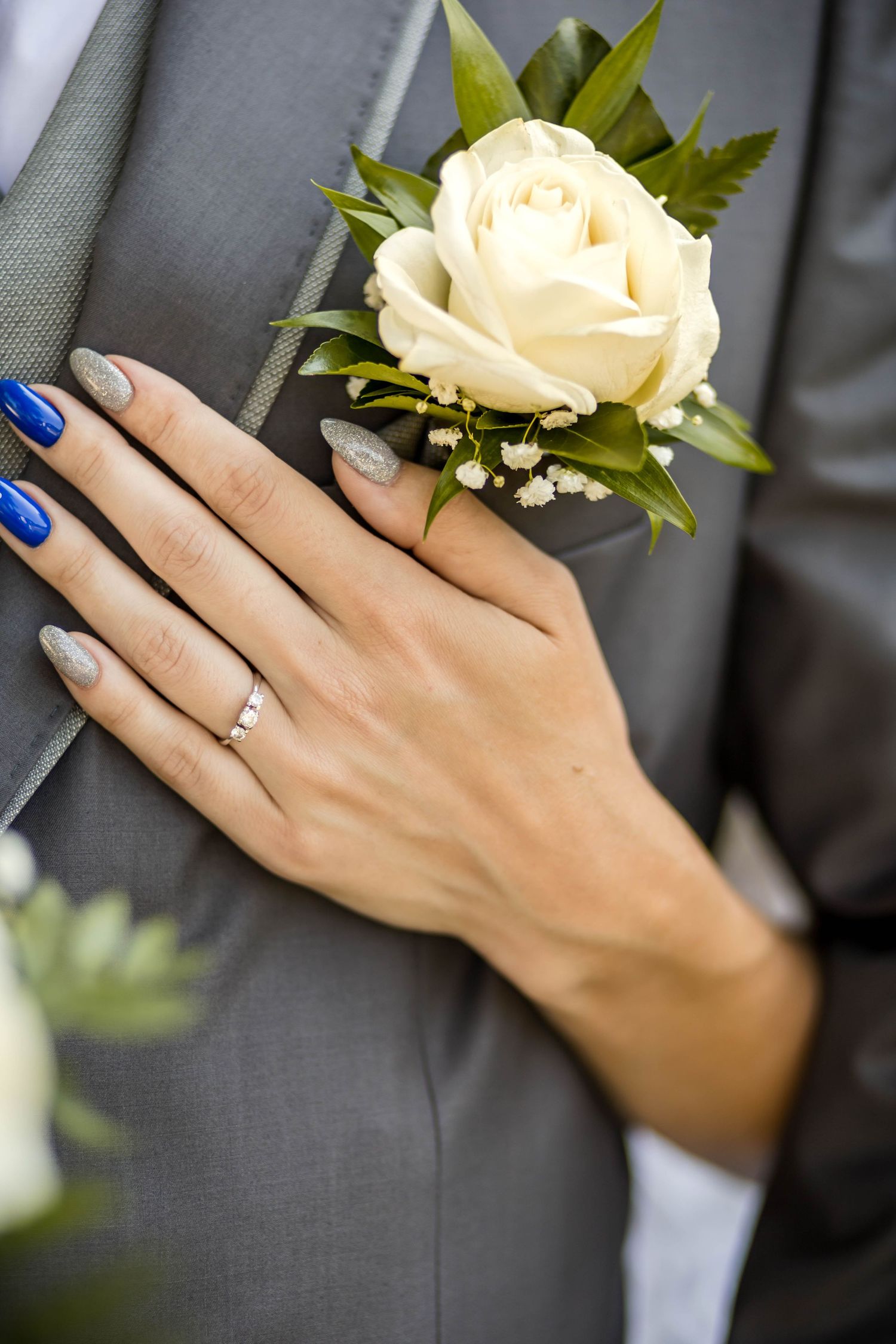 A woman's hand with blue nail polish and an engagement ring rests on a gray suit jacket with a white rose boutonniere.