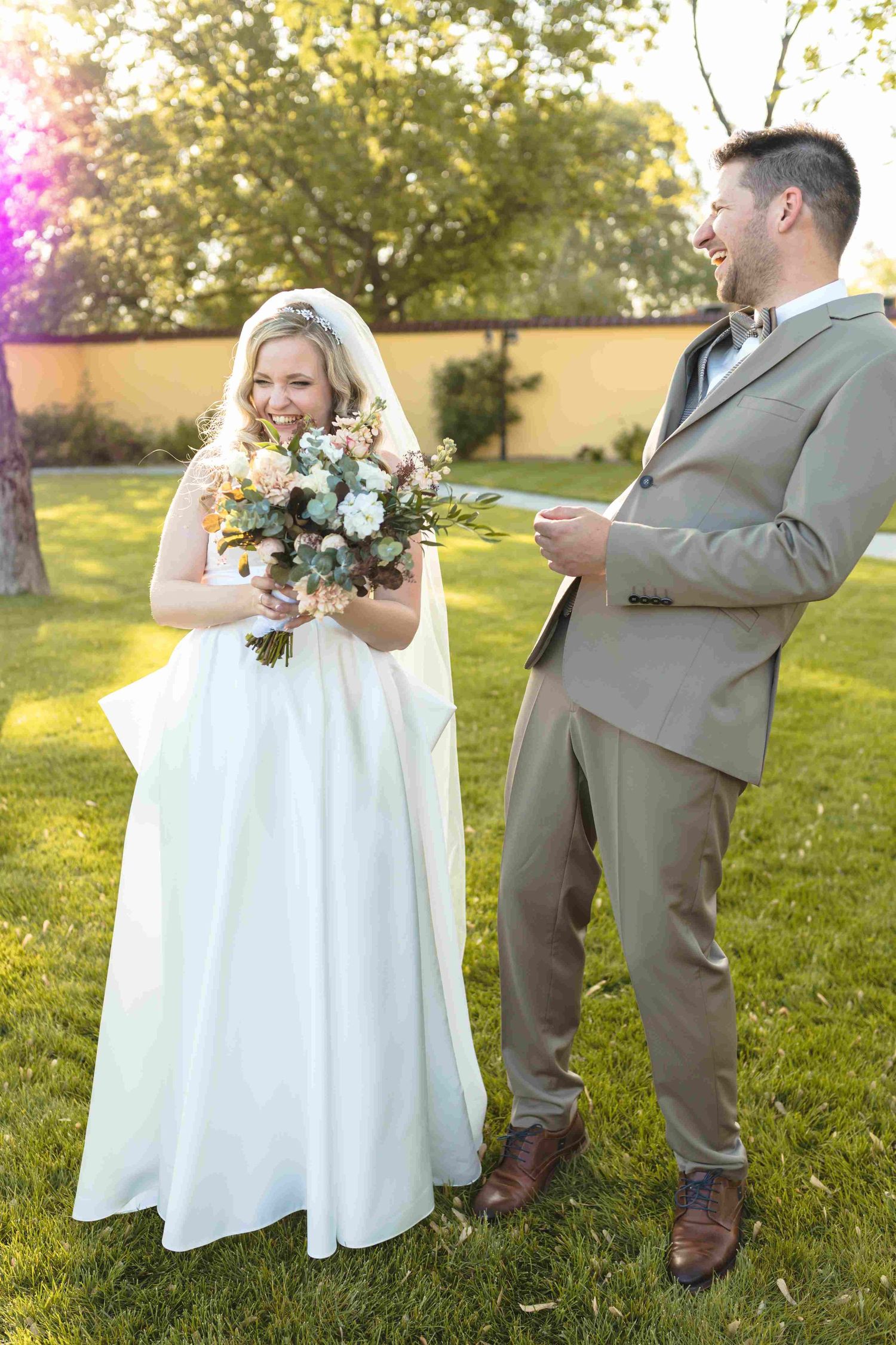 A bride in a white wedding dress holds a bouquet while standing with a groom in a tan suit on a sunny lawn.