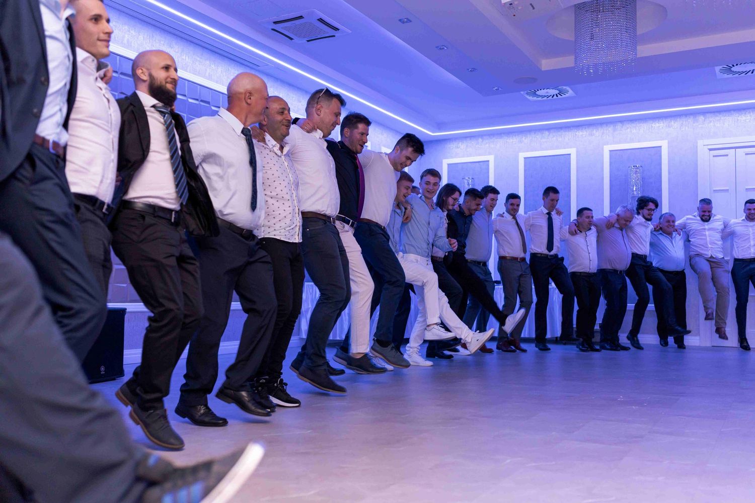 Dancers in white shirts and dark pants perform synchronized line dance moves in a blue-lit studio space.