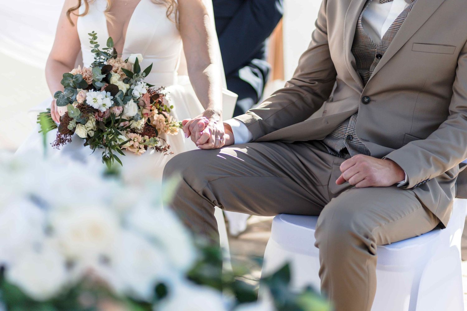 A couple holds hands while seated during an outdoor wedding ceremony with floral decorations in view.
