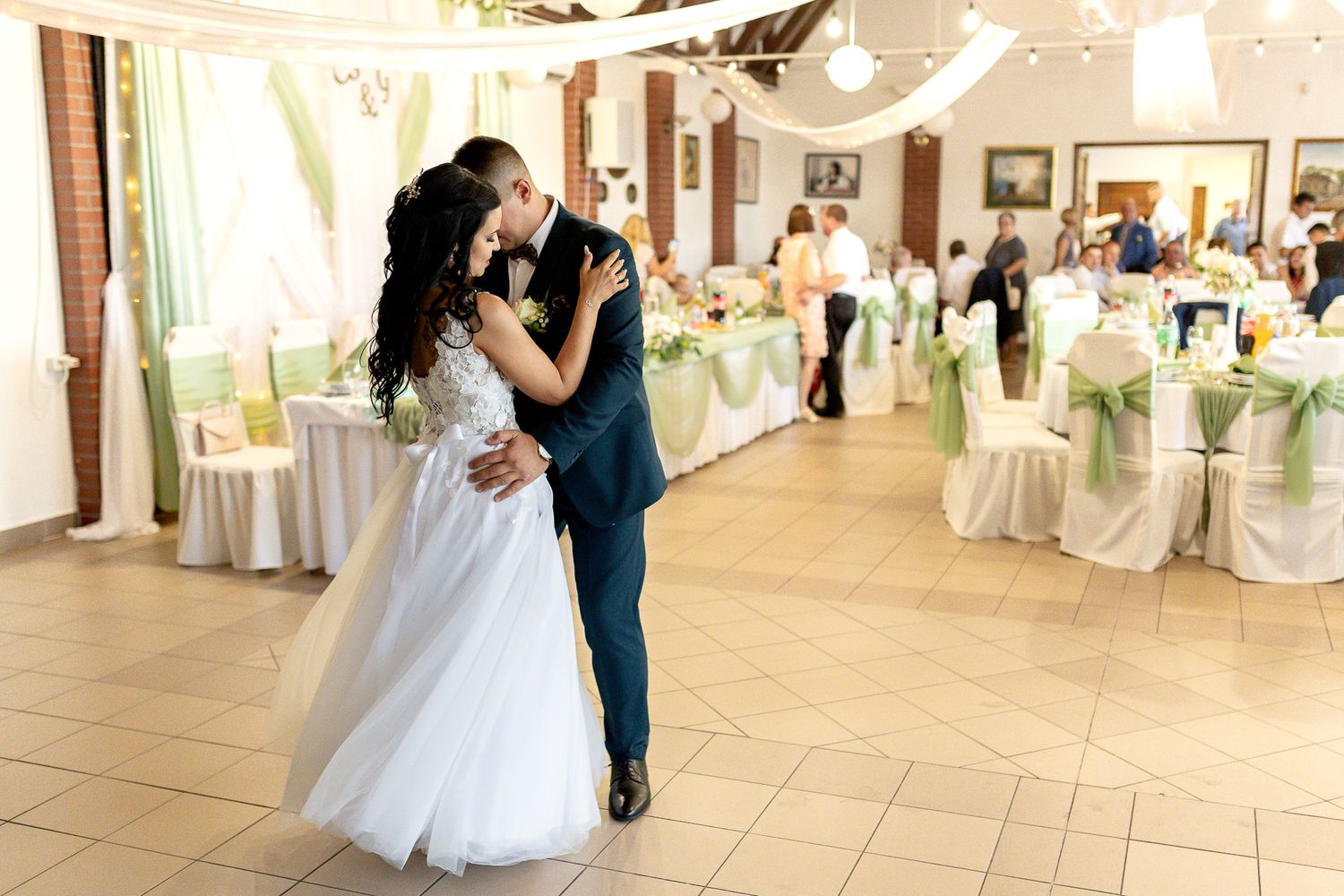 Elegant couple dancing in a white-decorated reception hall with round tables and chairs arranged for a wedding celebration.