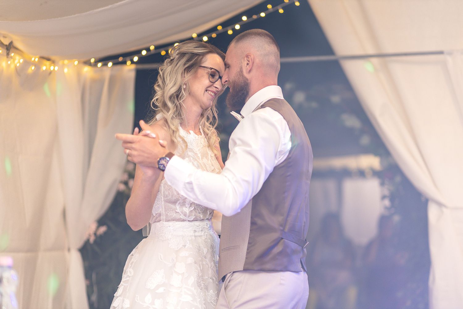 A couple shares their first dance at a wedding reception under twinkling string lights and white drapes.