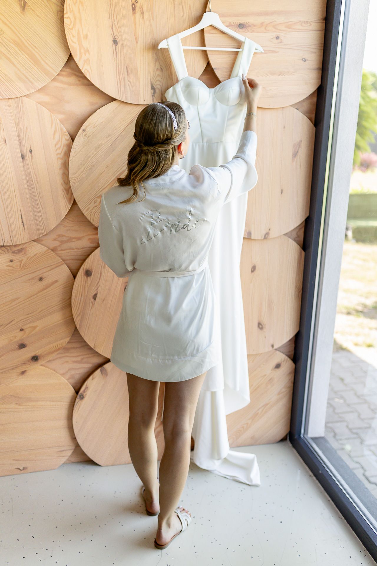 Someone in a white robe adjusts a wedding dress hanging on display against wooden wall decor.