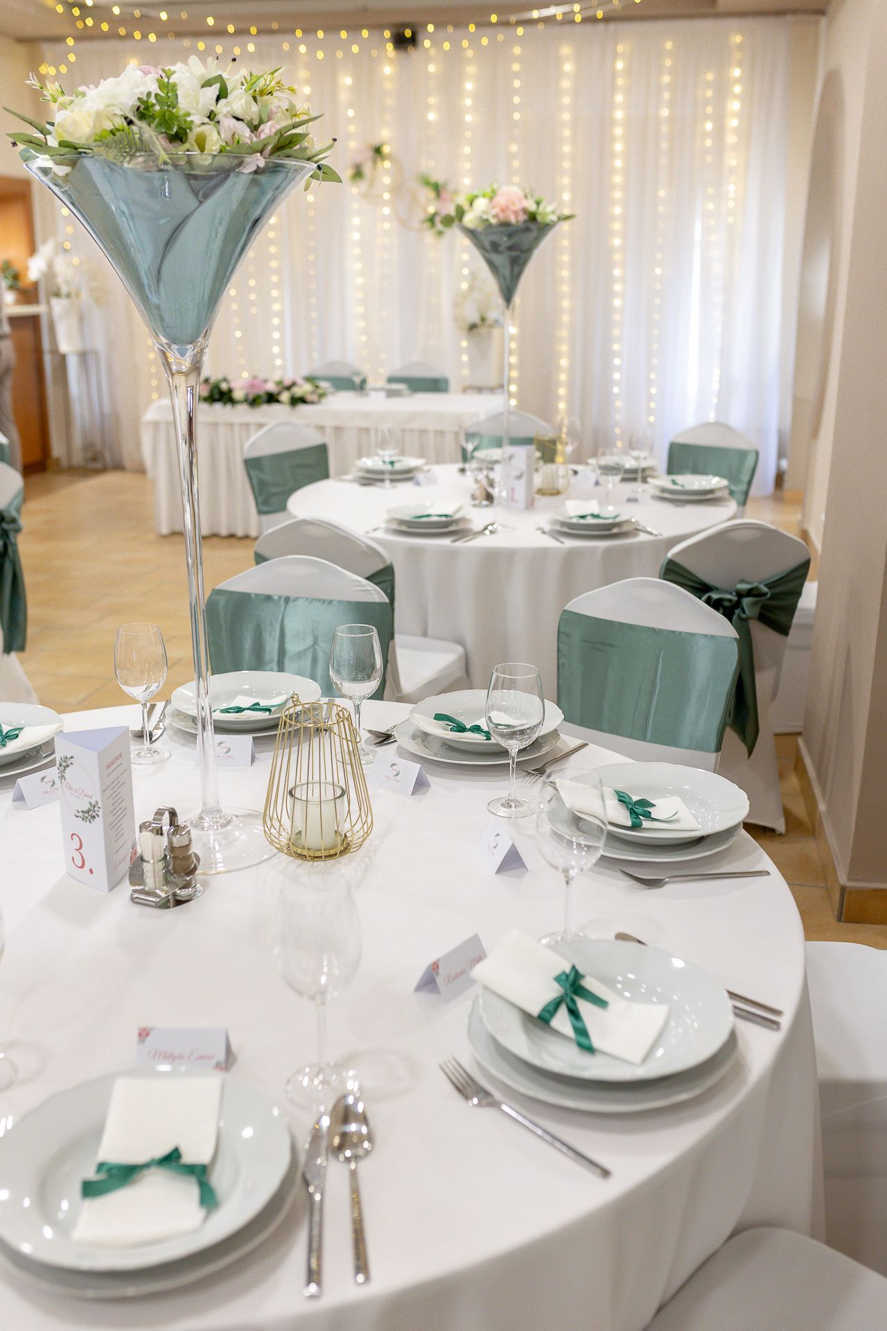 Elegant wedding reception tables decorated in white and turquoise with string lights and martini glass centerpieces.