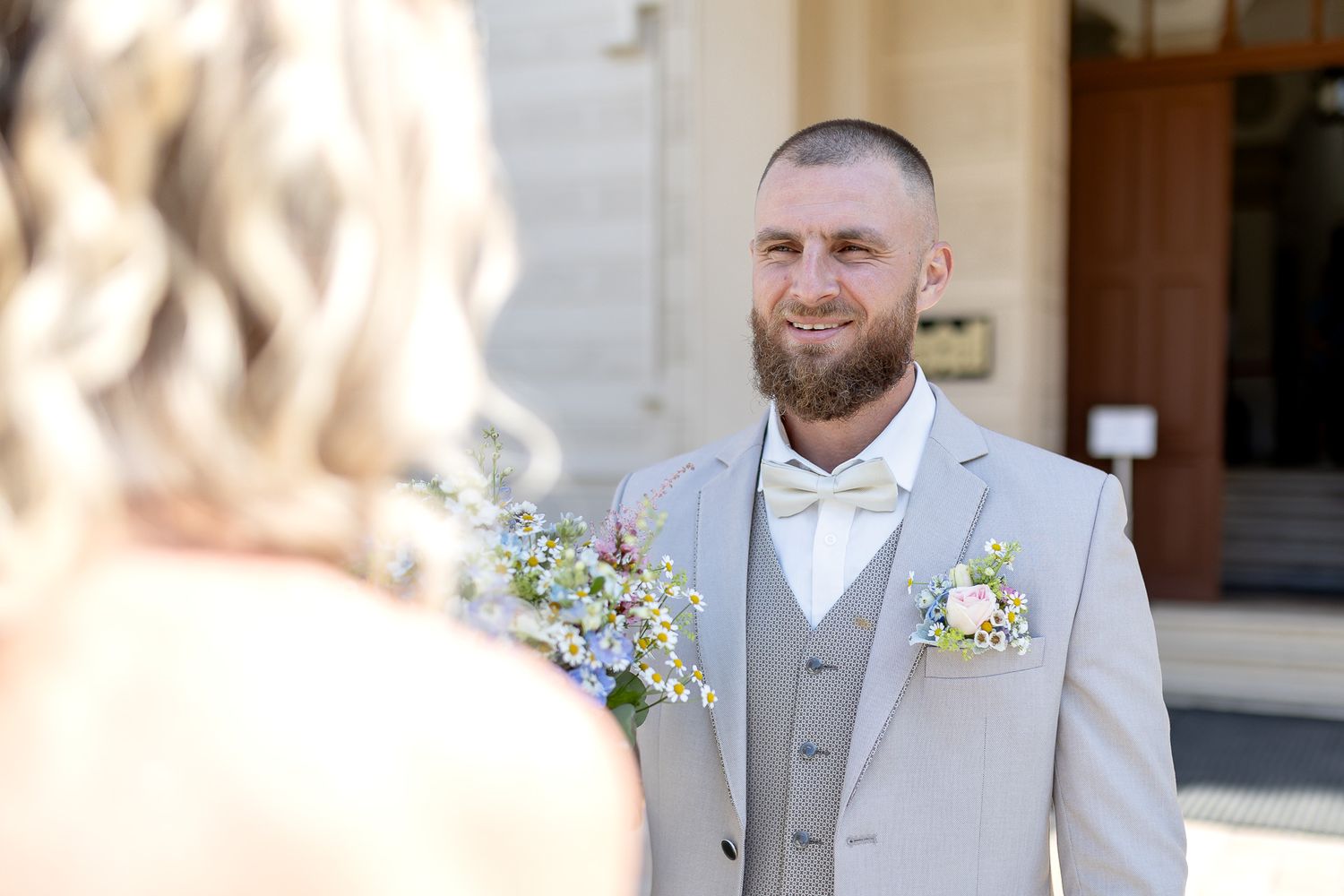 A groom in a light gray three-piece suit smiles while looking at his bride during their outdoor wedding ceremony.