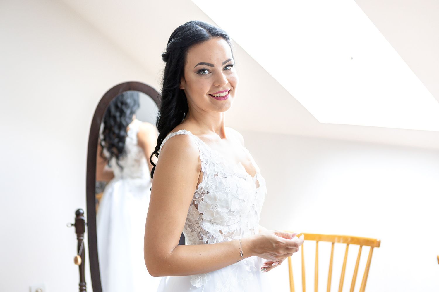 A bride in a white lace wedding dress stands by a window in soft natural light.