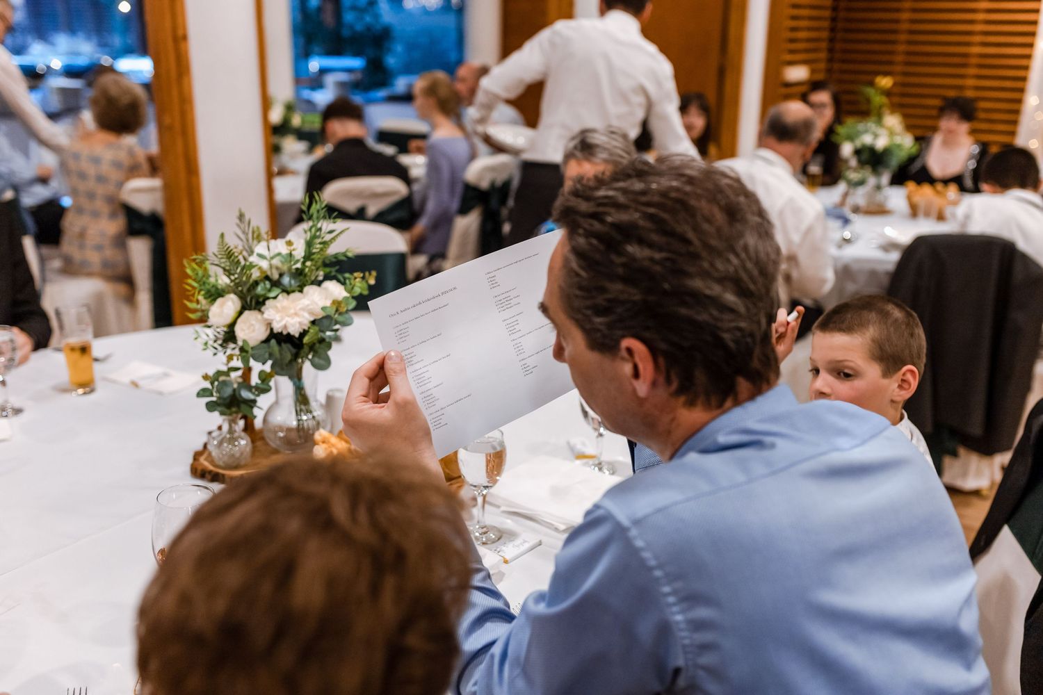 Wedding reception guests seated at round tables in an elegant banquet hall with white tablecloths and string lights overhead.