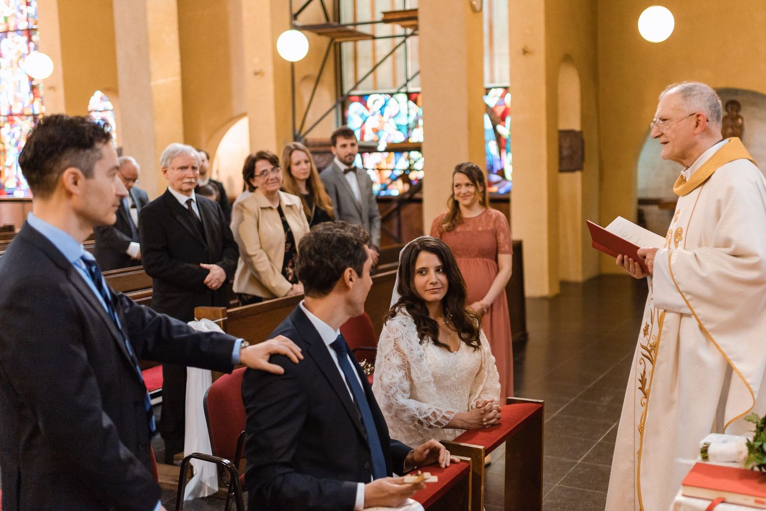 A group gathers in a church with stained glass windows and cream-colored walls during a religious ceremony.