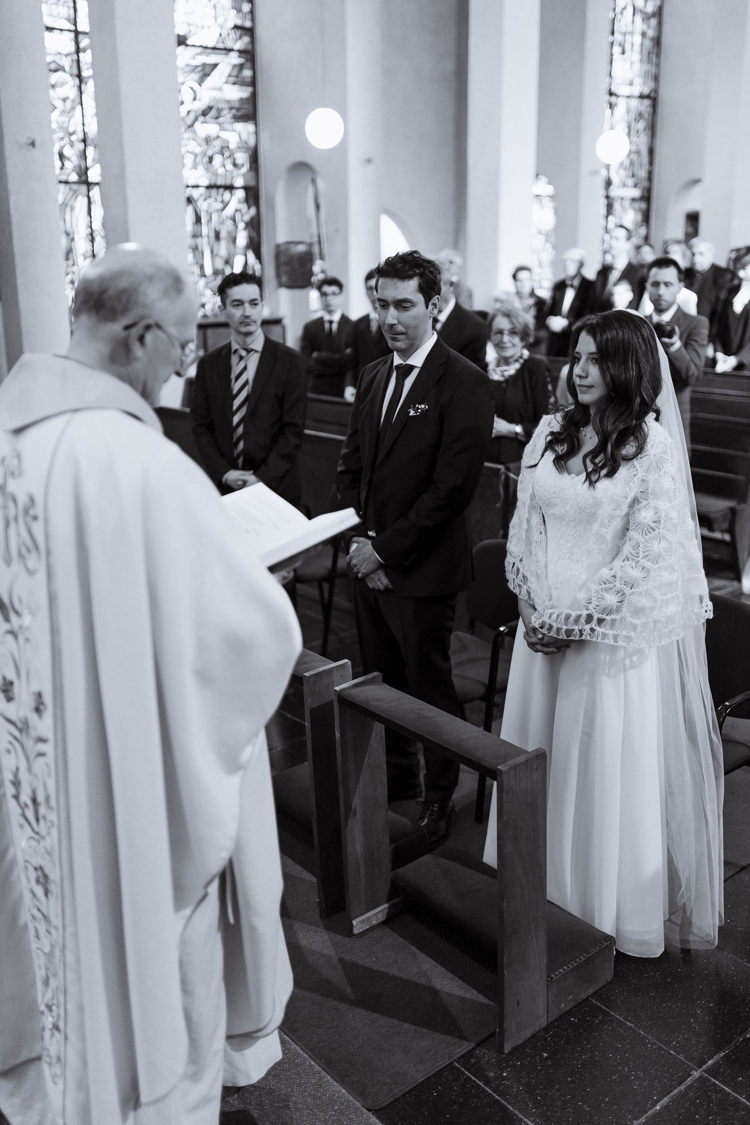 Black and white photo of church interior with clergy in white robes and guests seated in wooden pews during ceremony.