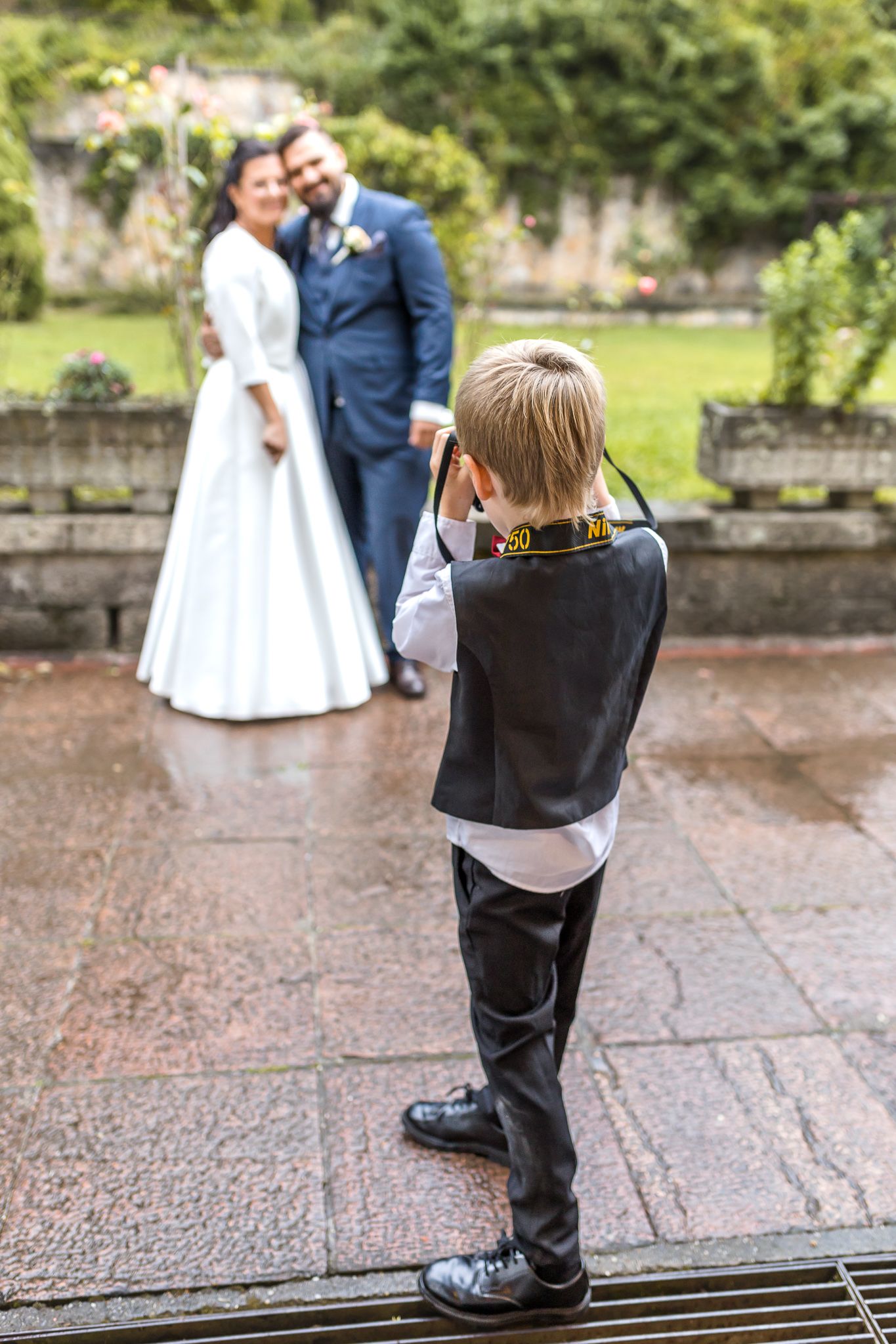 Young wedding photographer in formal attire crouches down to capture the perfect shot at an outdoor venue.