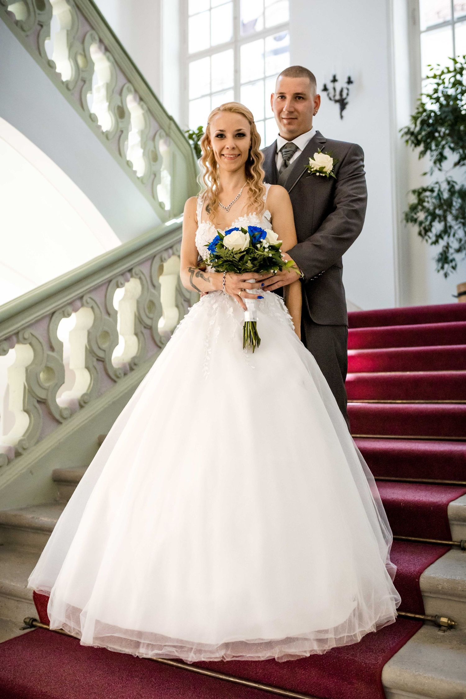 A wedding couple poses on elegant stairs with red carpet and ornate white railings in a grand interior setting.