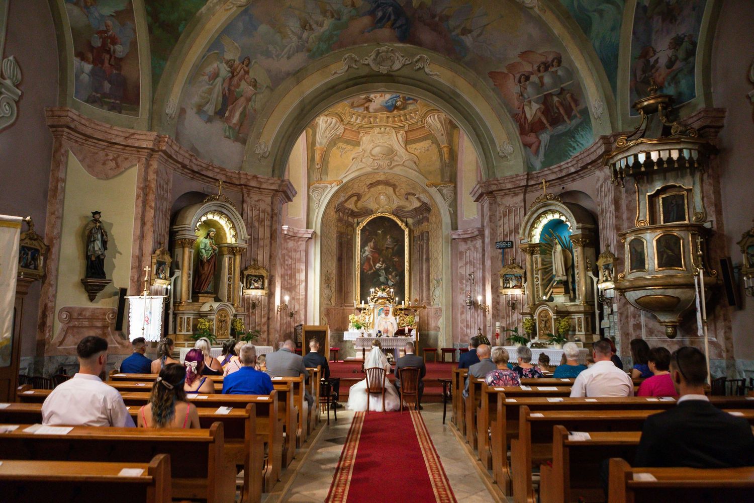 Interior view of an ornate Catholic church with painted ceilings, arched columns, and pews filled with worshippers.