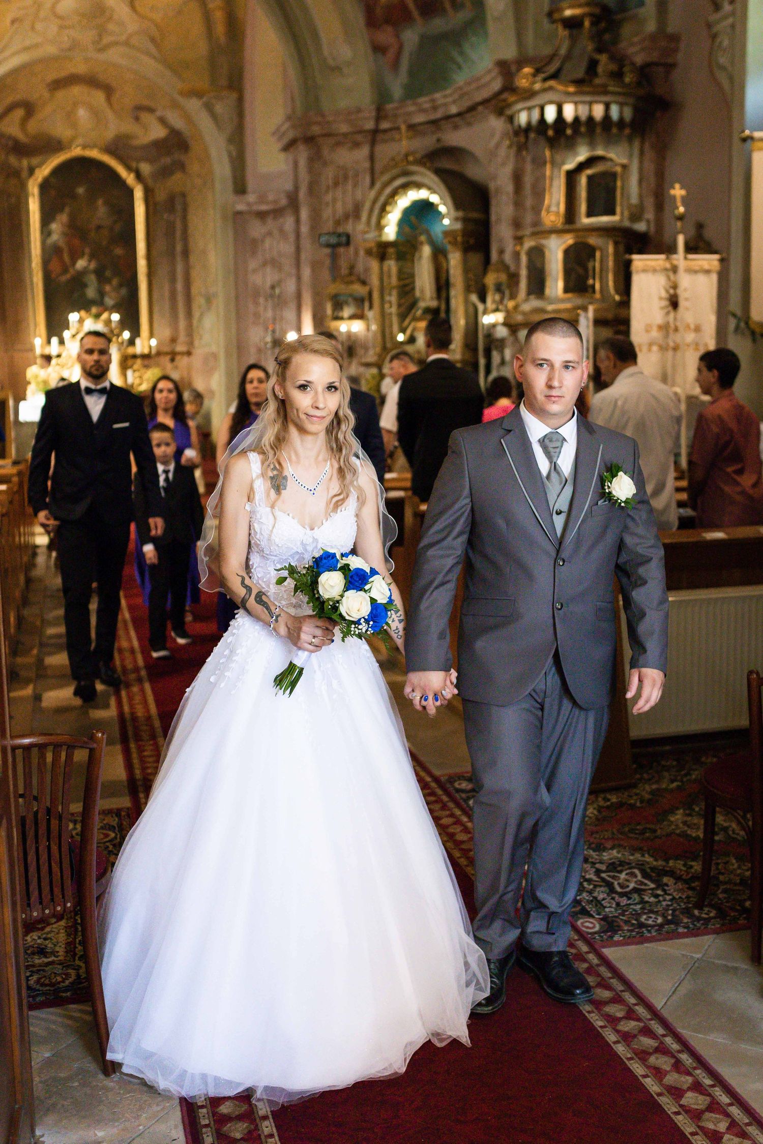 Wedding couple walks down ornate church aisle with red carpet and golden decor in traditional ceremony setting.