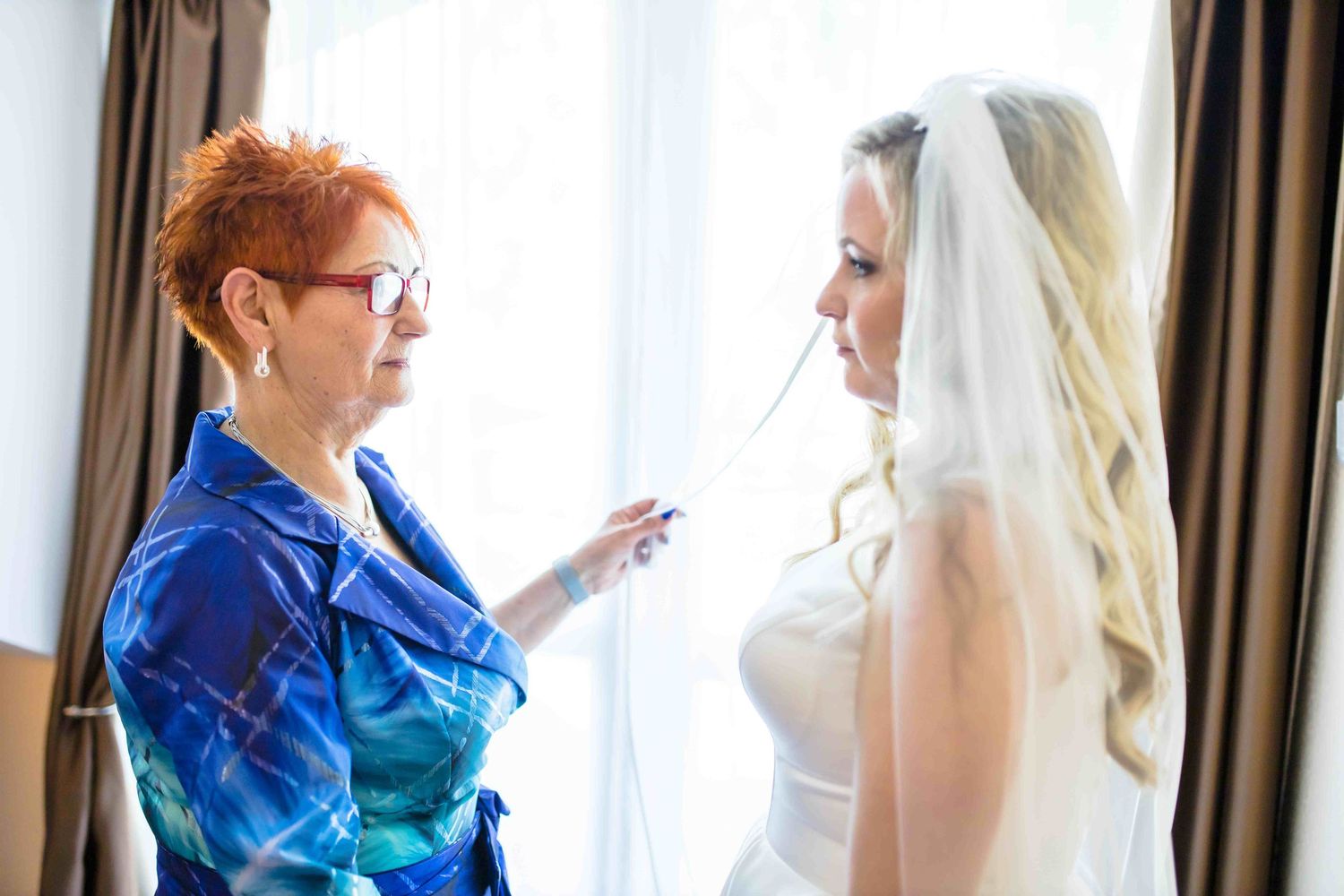 A bride in a white wedding gown and veil stands by a window during wedding preparations.