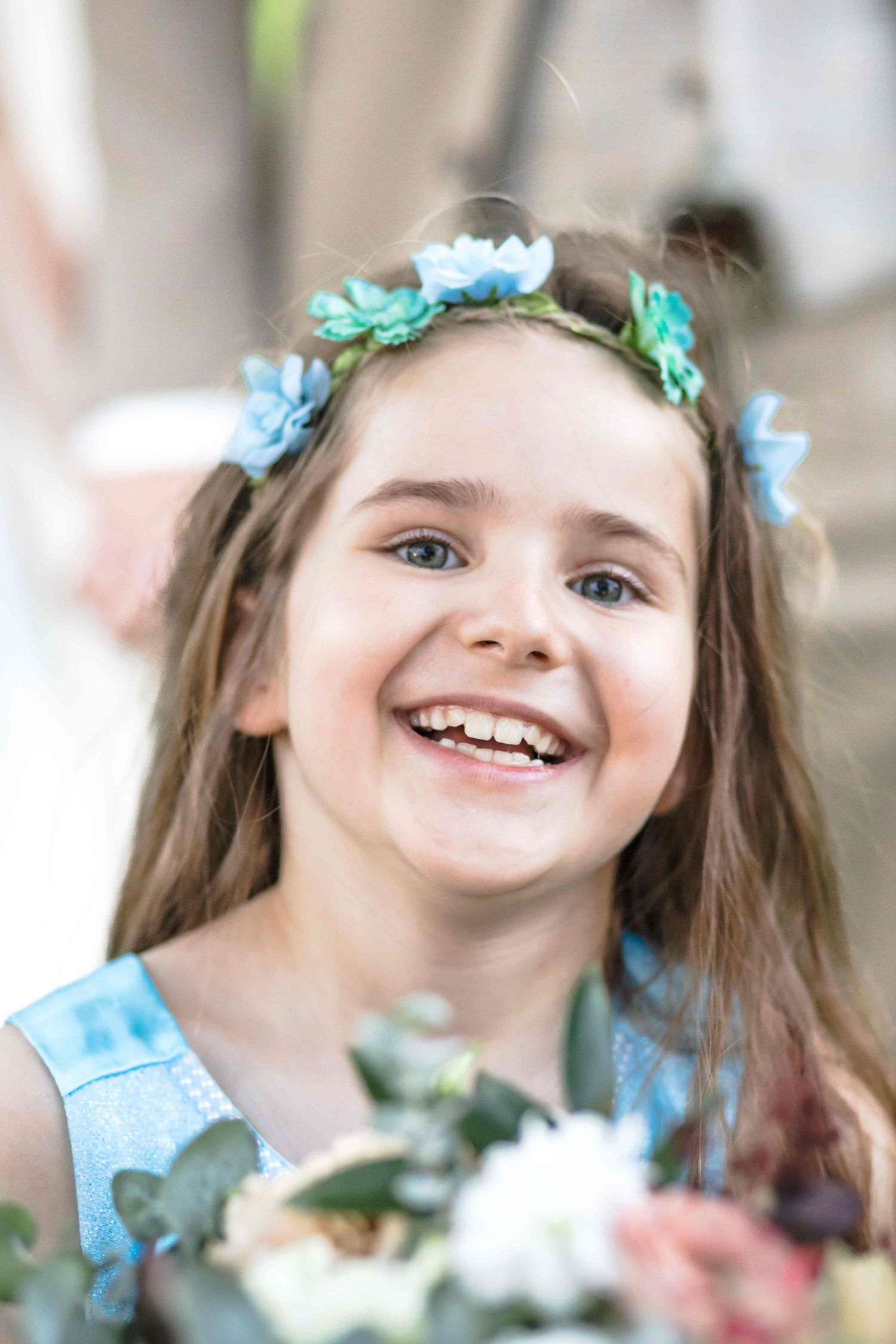 A joyful child wearing a blue dress and floral crown smiles brightly in a soft-focus portrait.