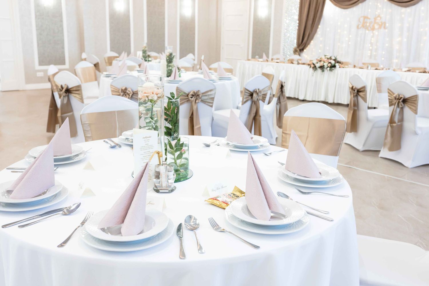 Elegant wedding reception tables set with white linens, blush napkins, and delicate floral centerpieces in a bright venue.