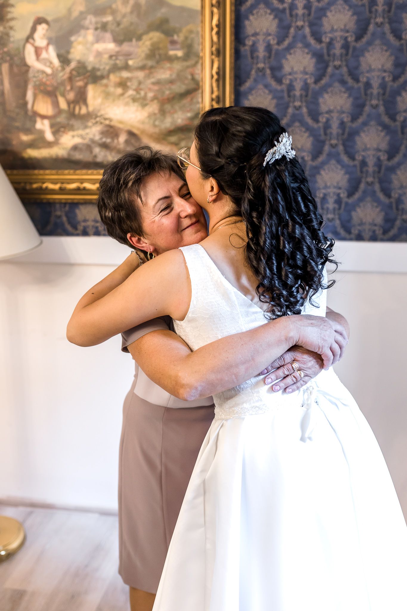 Two people share a tender embrace in front of an ornate blue wallpapered wall with gold-framed artwork.