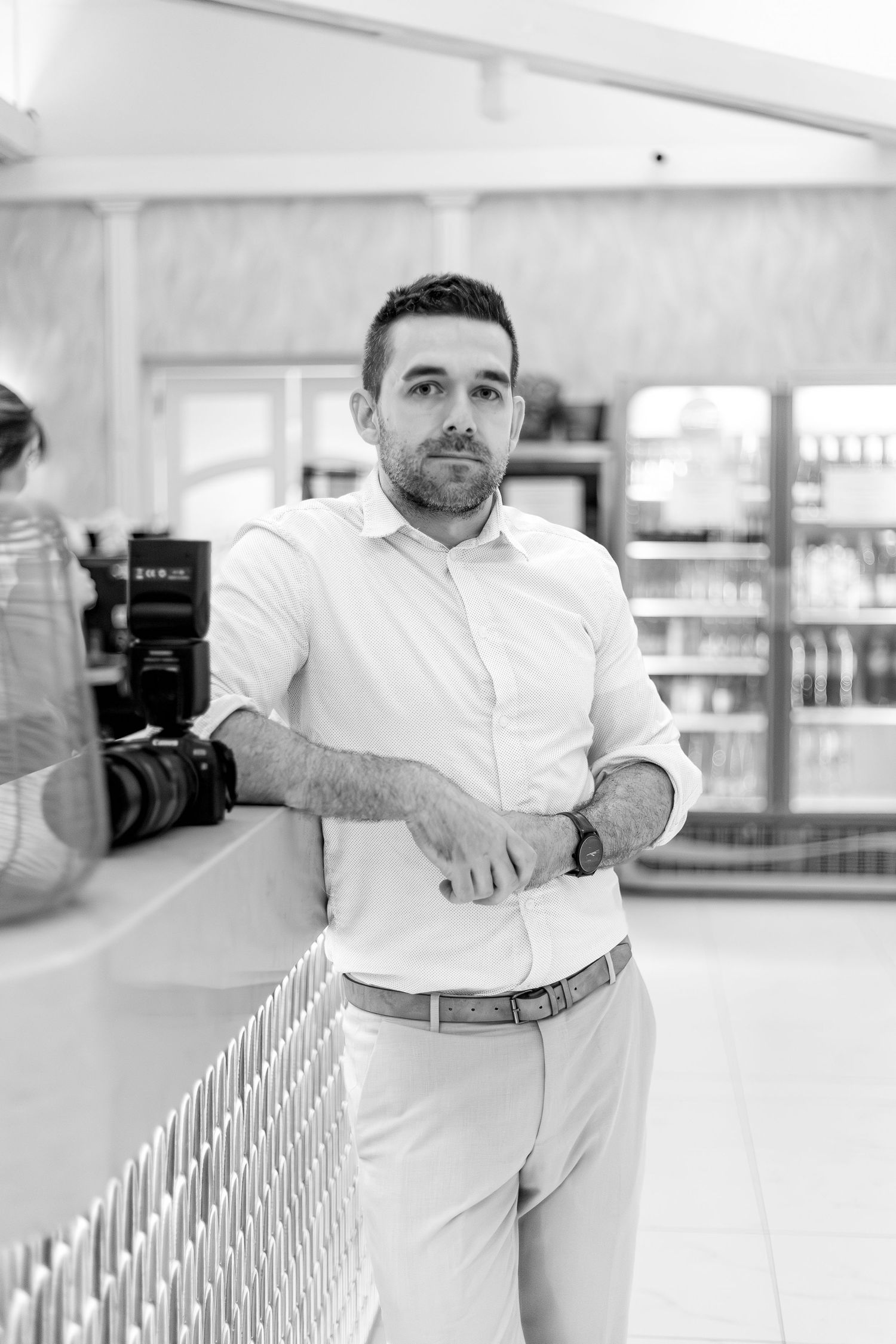 Professional photographer in white button-down shirt and khaki pants leans against railing in bright indoor setting.