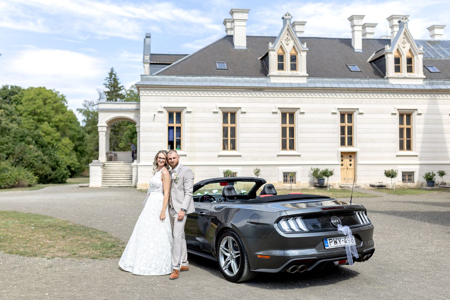 A wedding couple poses next to a silver sports car outside an elegant white mansion.