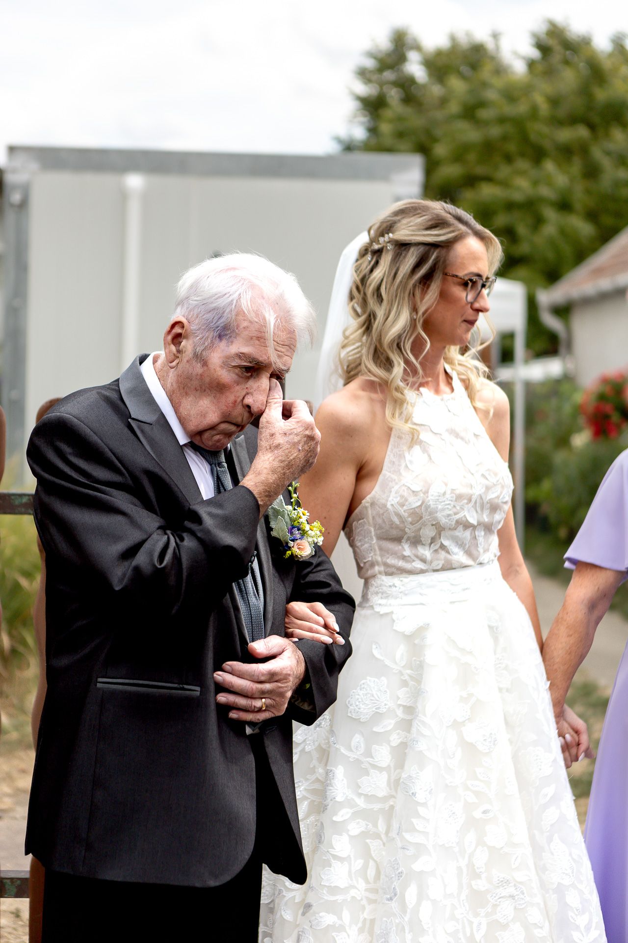 A touching moment at a wedding as an elderly man in a black suit kisses a bride's hand in an outdoor setting.