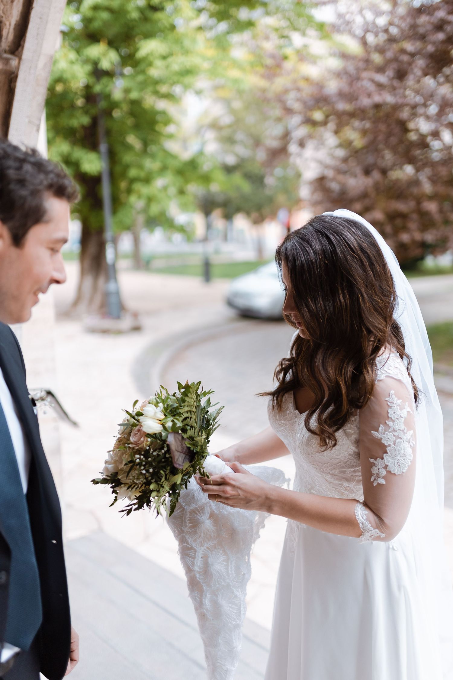 A bride in a white lace dress looks at her phone while holding a bouquet outdoors.