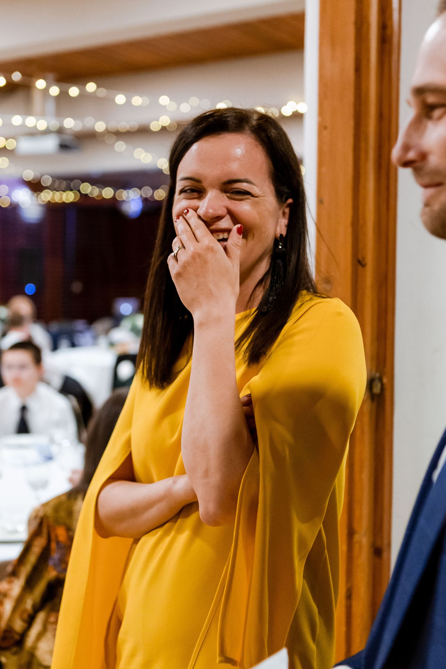 Woman in bright yellow sweater laughs joyfully during an indoor event with twinkling lights in background.