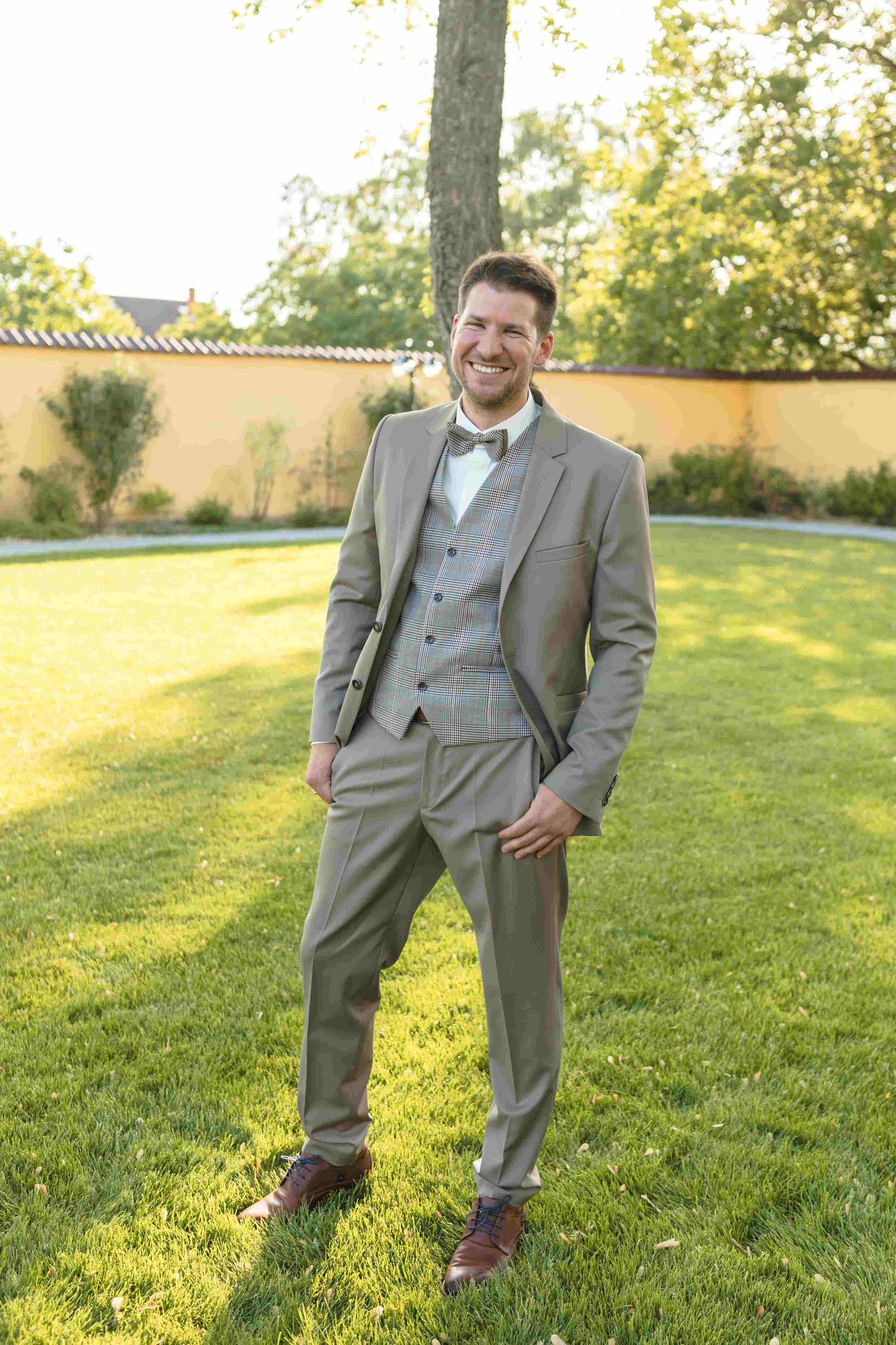 A groom in a light gray three-piece suit with bow tie stands smiling on a sunny lawn during a wedding.