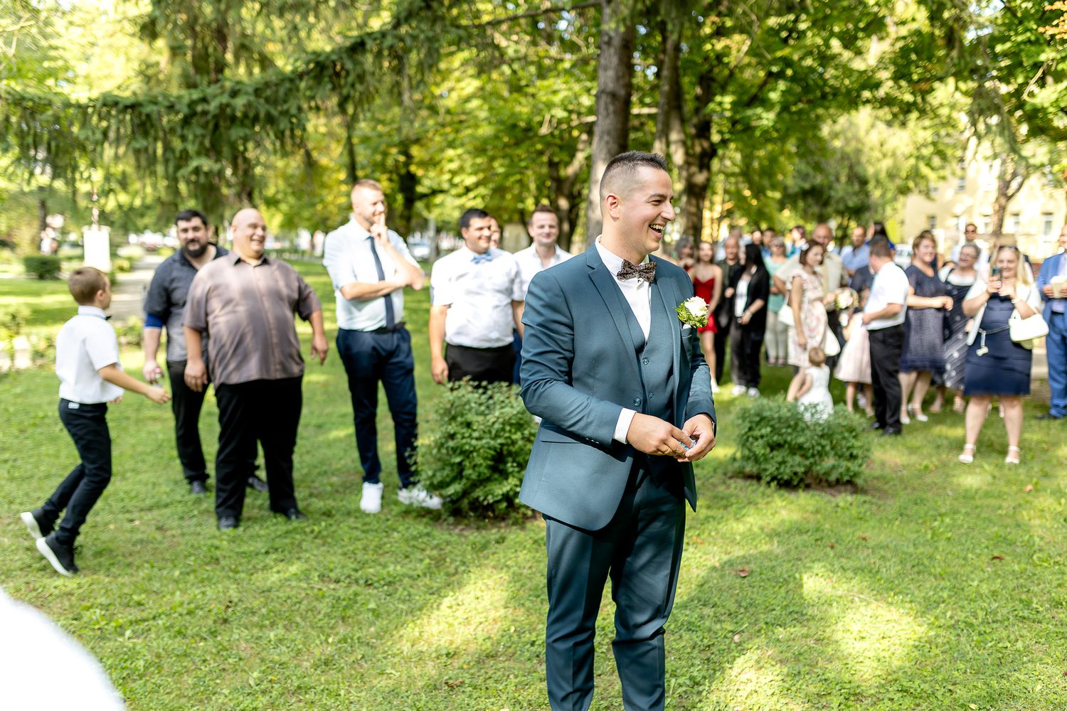 A group of wedding guests dancing and mingling outdoors in a sunny garden setting with lush green trees.