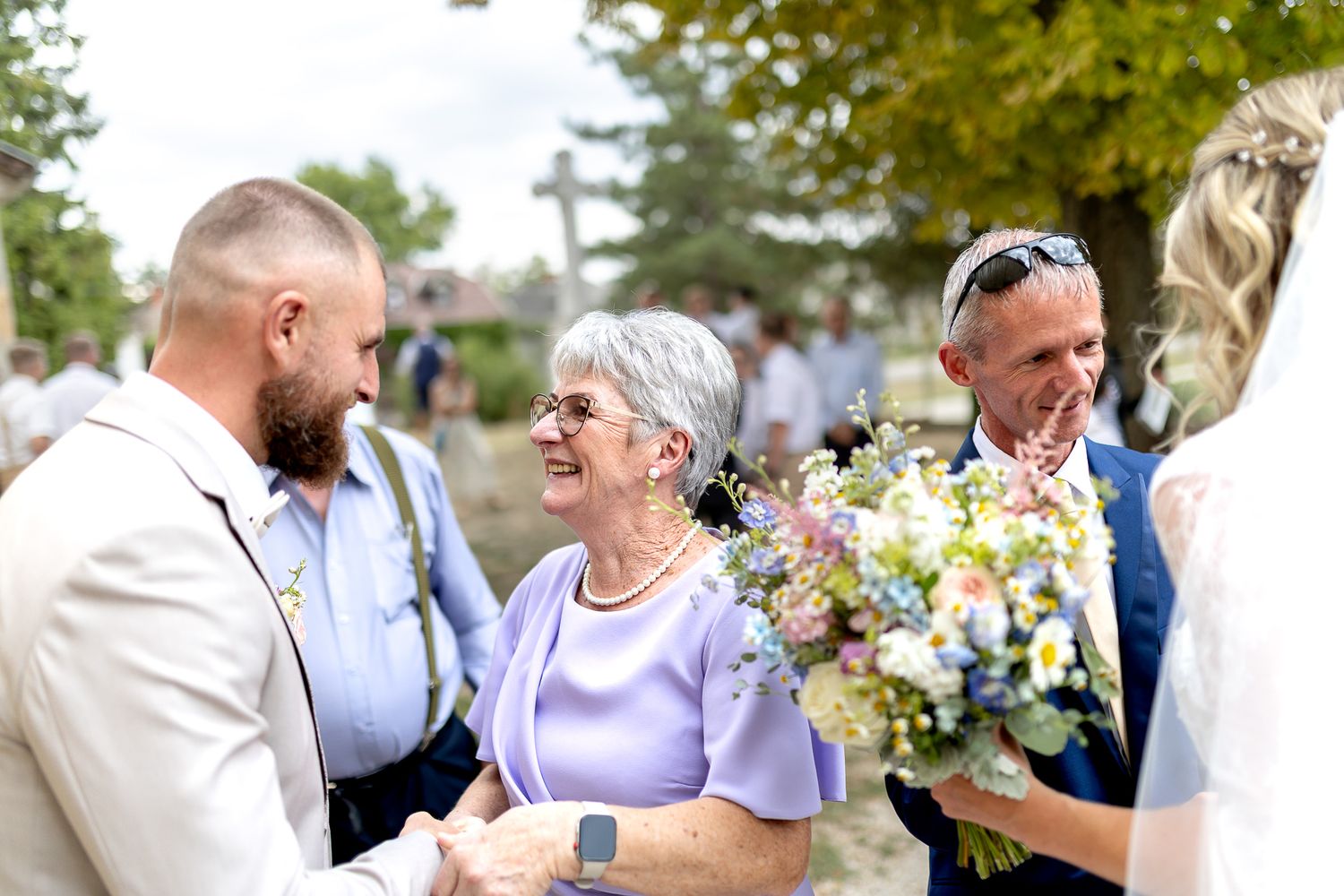 Wedding guests gather outdoors on a sunny day with colorful bouquet of flowers and festive summer attire.