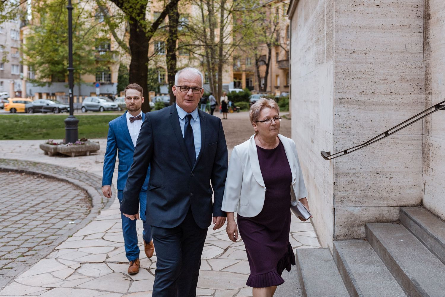 A group of people in formal attire walk together along a stone building exterior during daytime.