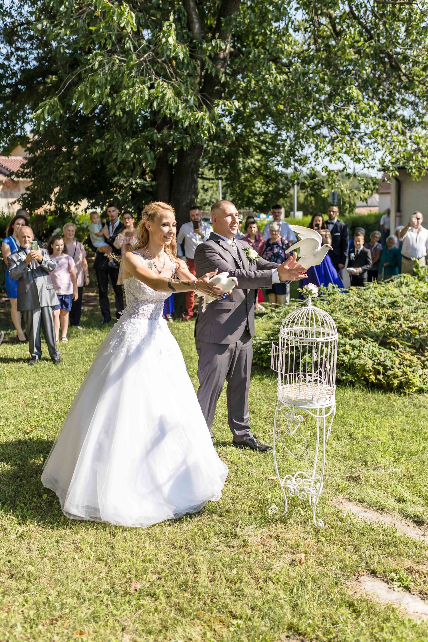 A couple in wedding attire releases doves during an outdoor garden ceremony as guests look on.