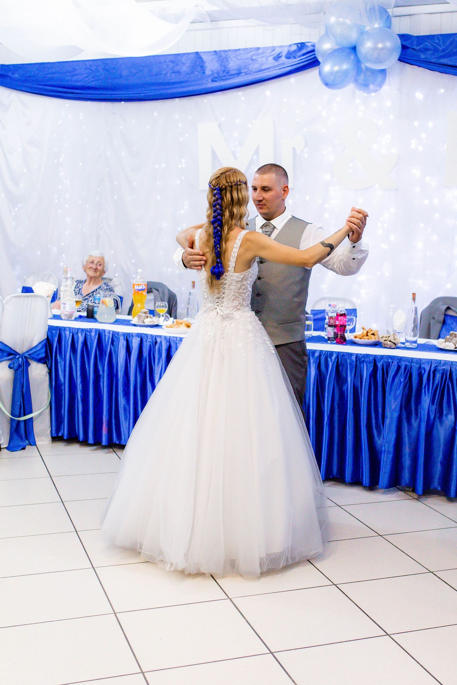 Couple shares their first dance at wedding reception with royal blue decor and white tablecloths in background.