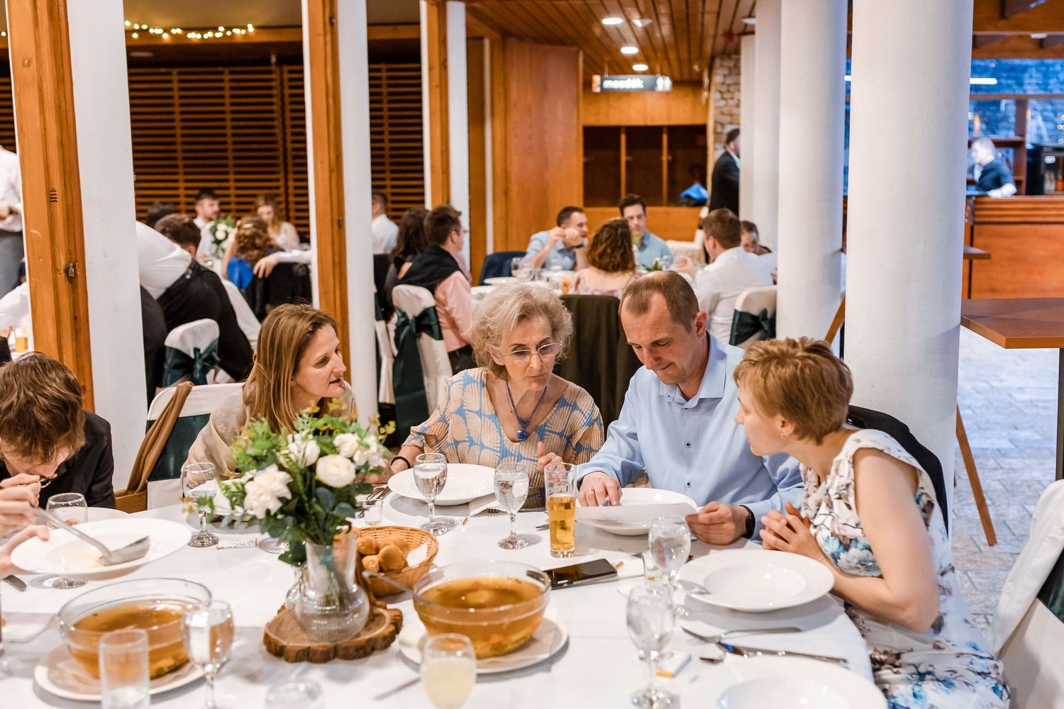 Diners seated at a long wooden table in a restaurant enjoy a meal together with plates and drinks spread across the surface.